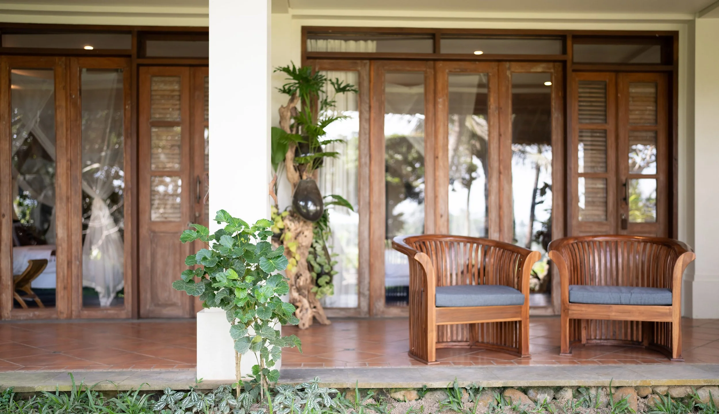 The image shows a porch or patio area with wooden sliding doors, green houseplants, and wooden chairs with blue cushions. High-end retreat center in Pejeng, Ubud, Bali