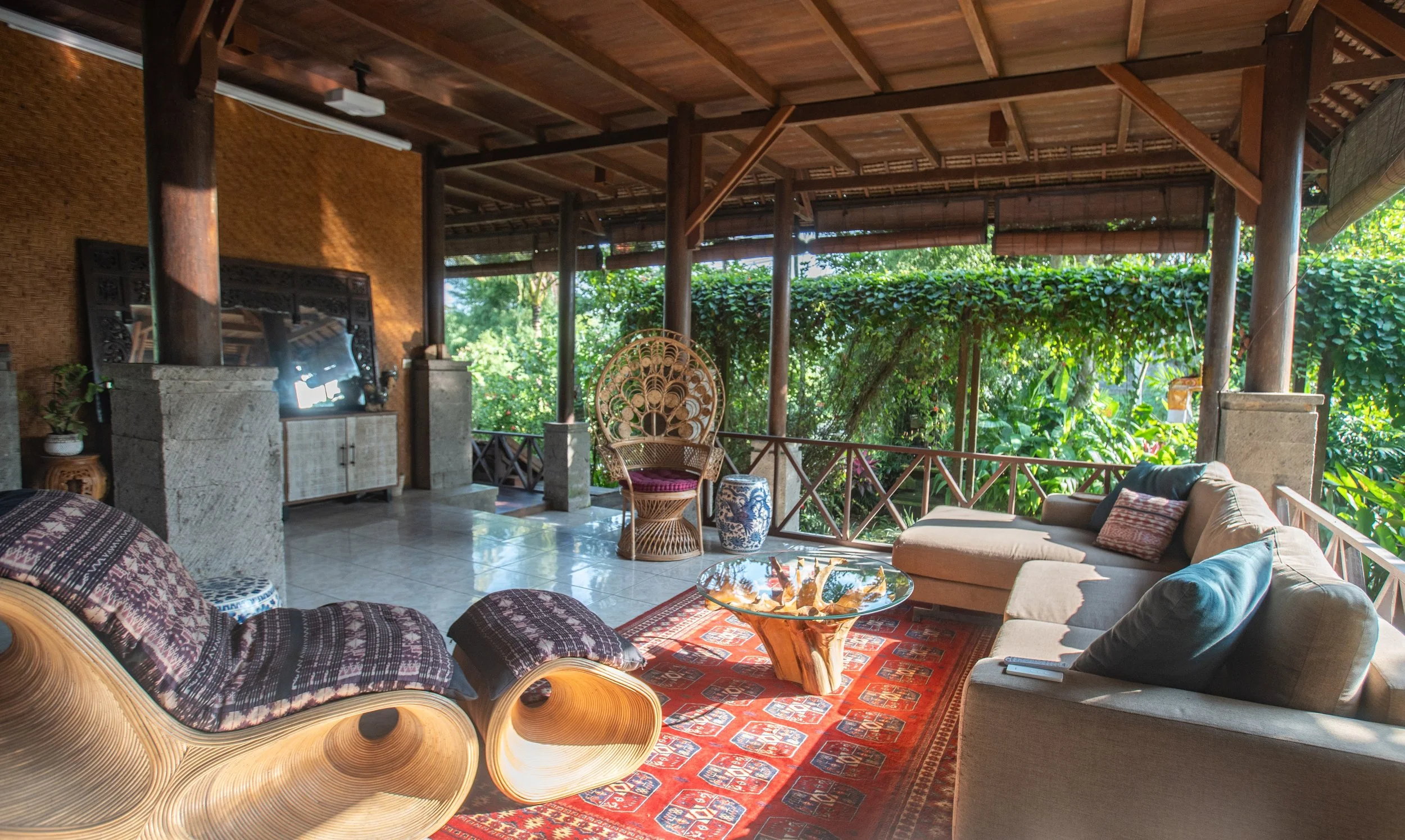 Living room with wooden ceiling and lush green view outside, featuring a beige sofa with pillows, a glass coffee table, woven chairs, a patterned rug, and decorative elements.