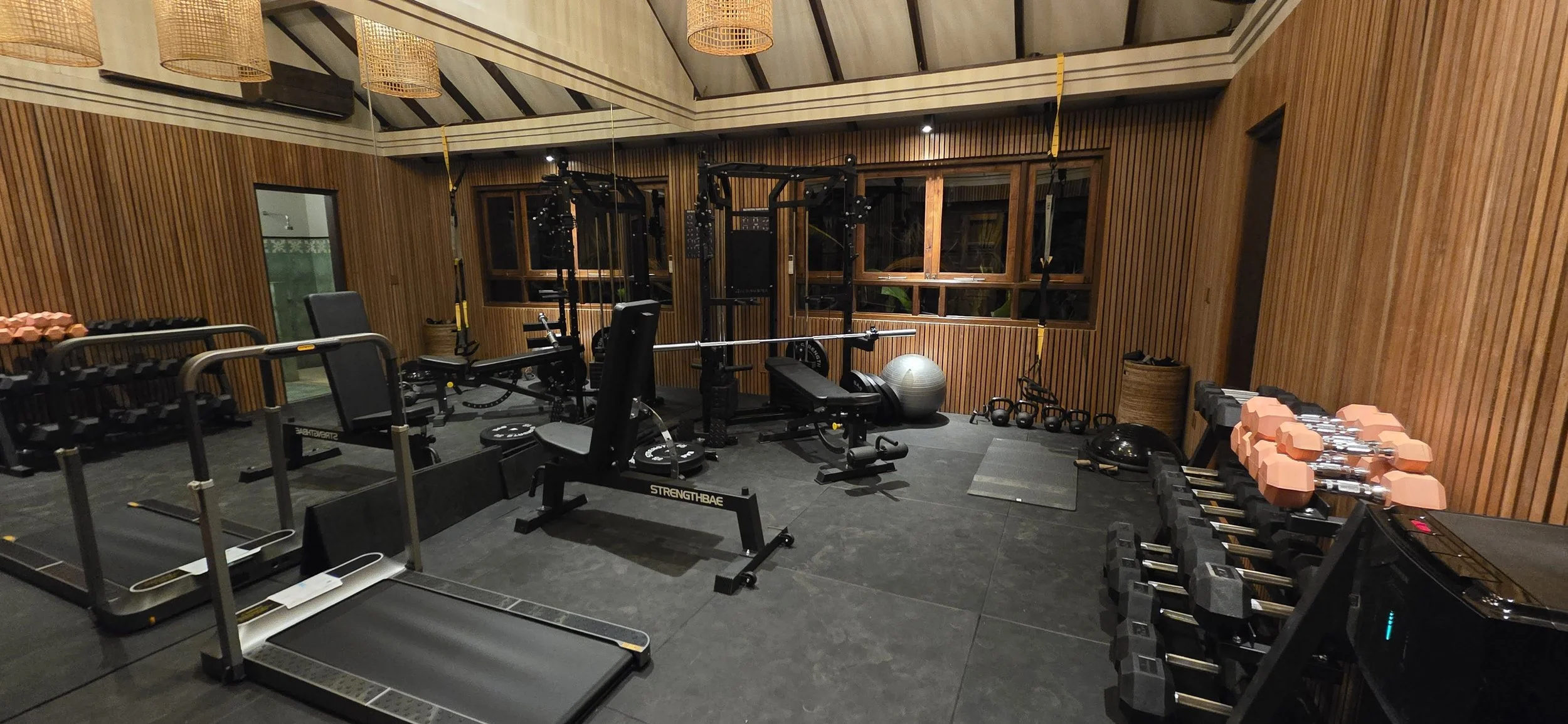A home gym with wooden walls, containing exercise equipment including dumbbells, kettlebells, a treadmill, weight benches, resistance bands, an exercise ball, and a medicine ball, under warm lighting. High-end retreat center in Pejeng, Ubud, Bali