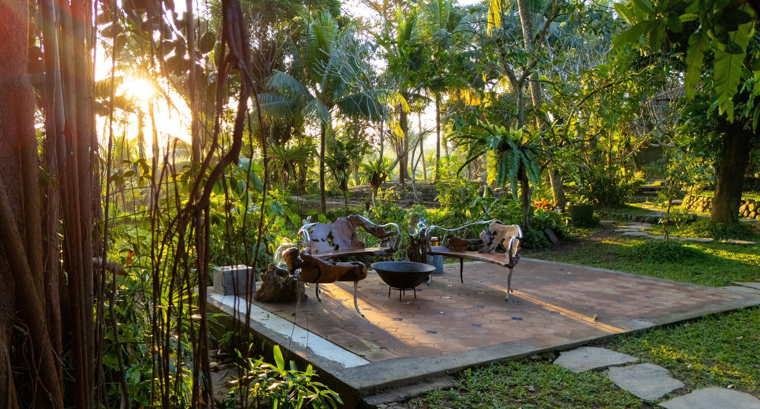 A garden patio area with wooden chairs, a round fire pit, and tall greenery including trees and plants, illuminated by sunlight. High-end retreat center in Pejeng, Ubud, Bali