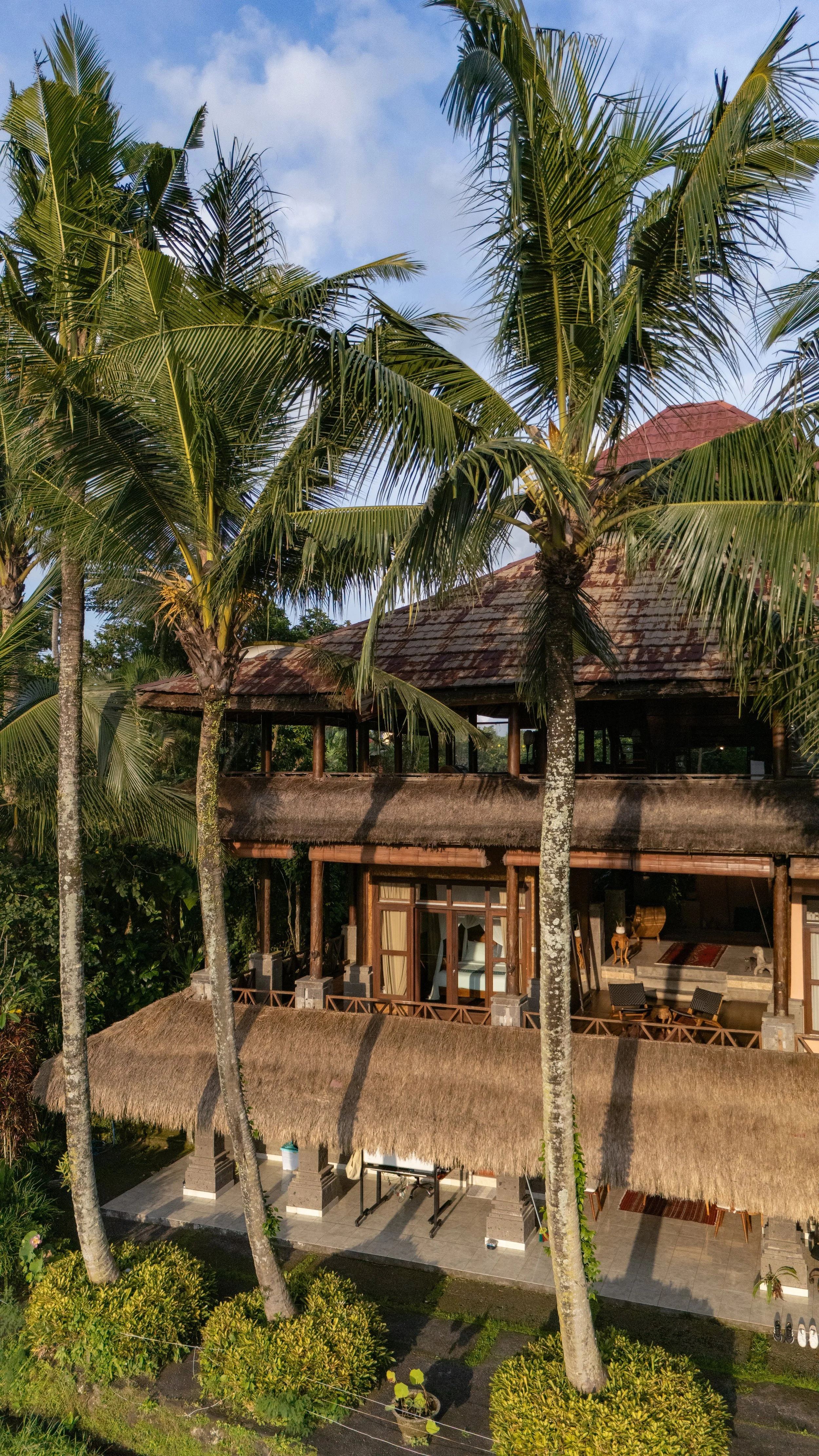 A tropical scene with tall palm trees in front of a house with a thatched roof and open patio area, under a clear blue sky. High-end retreat center in Pejeng, Ubud, Bali
