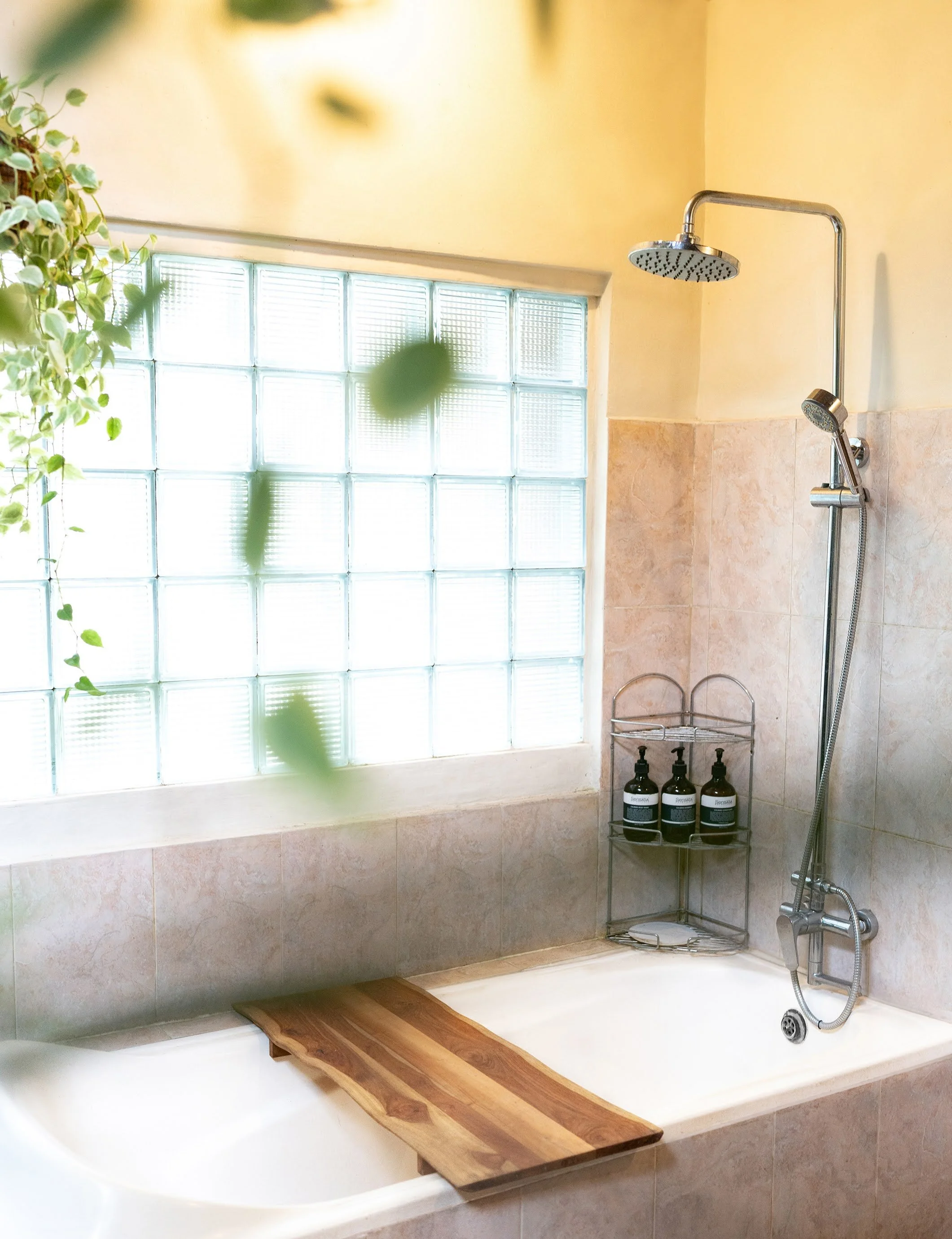 A bathroom shower and bathtub area featuring a frosted glass block window, a rain showerhead, a handheld showerhead, a small metal shelf holding three bottles, and a wooden bath tray across the tub.