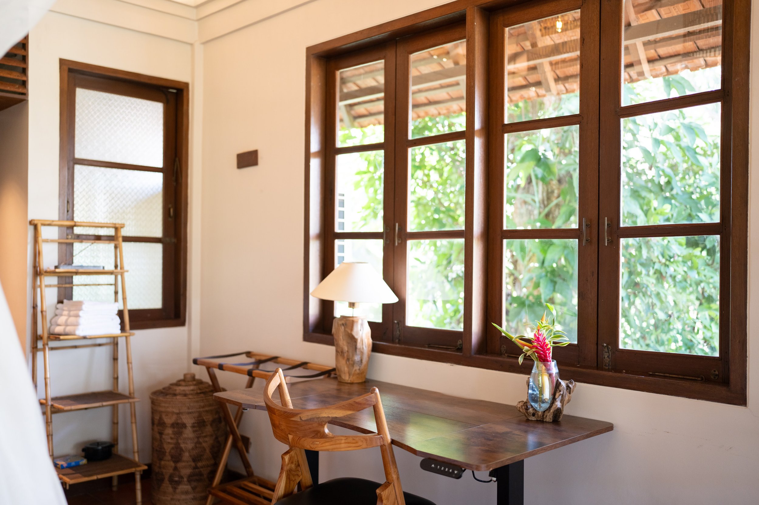 A cozy room with large wooden-framed windows showing green foliage outside. The room has a wooden table with a small colorful flower arrangement, a modern wooden chair, a lamp, and a shelf with towels and books. High-end retreat center in Pejeng,