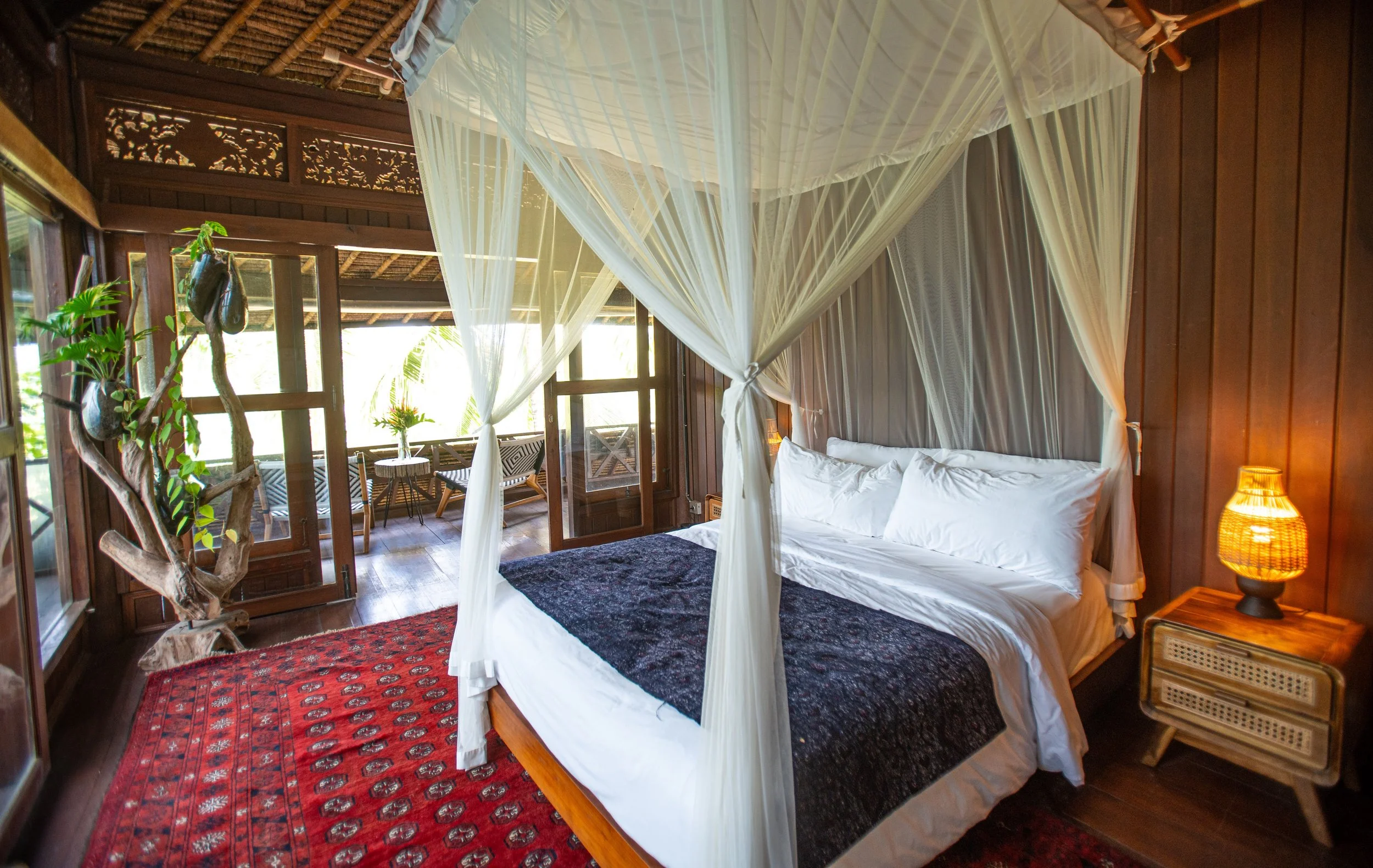 A cozy bedroom with a four-poster bed draped in white mosquito netting, wooden walls, a red patterned rug, a bedside lamp, a wooden side table, wicker chairs on a porch outside, potted plants, and large windows letting in natural light.