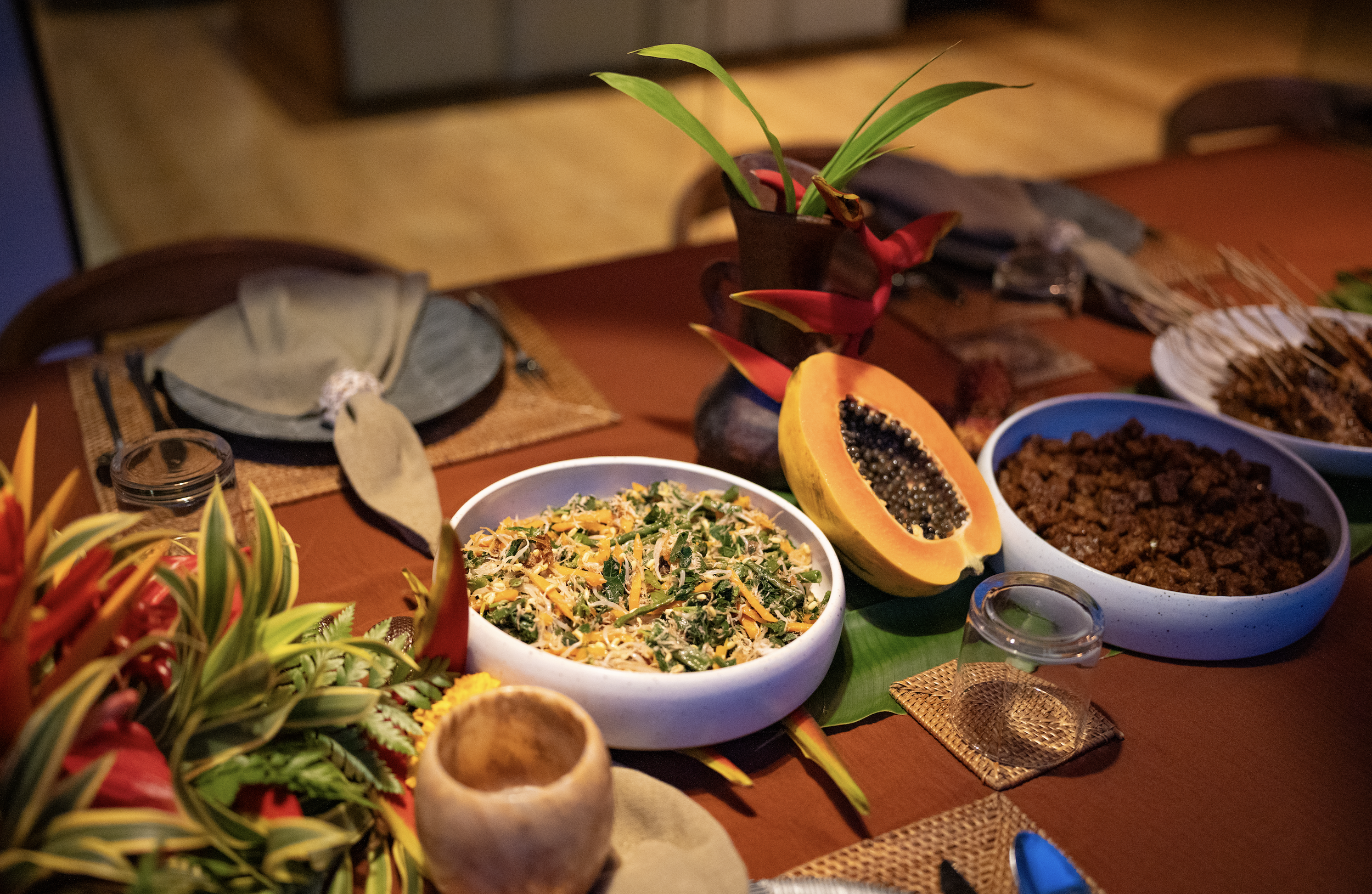 A table with a variety of food dishes, including a bowl of salad, a halved papaya, and assorted meats, surrounded by decorative flowers and tableware.