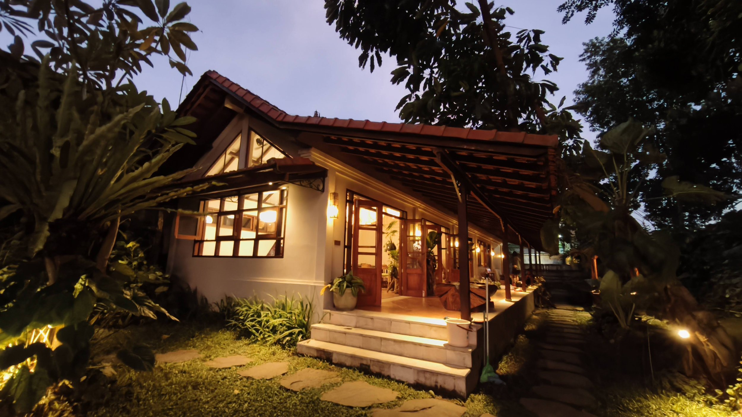A house with warm interior lighting, surrounded by trees and greenery, in the evening or night. High-end retreat center in Pejeng, Ubud, Bali