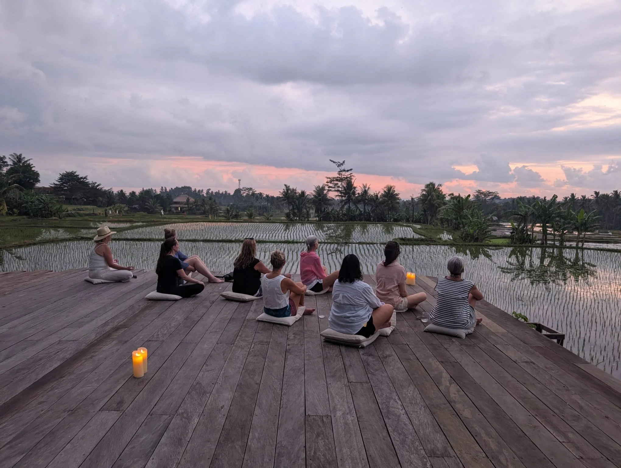 People sitting on cushions on a wooden deck facing a rice paddy field, with trees and cloudy sky in the background during sunset. A candle is placed on the deck. High-end retreat center in Pejeng, Ubud, Bali