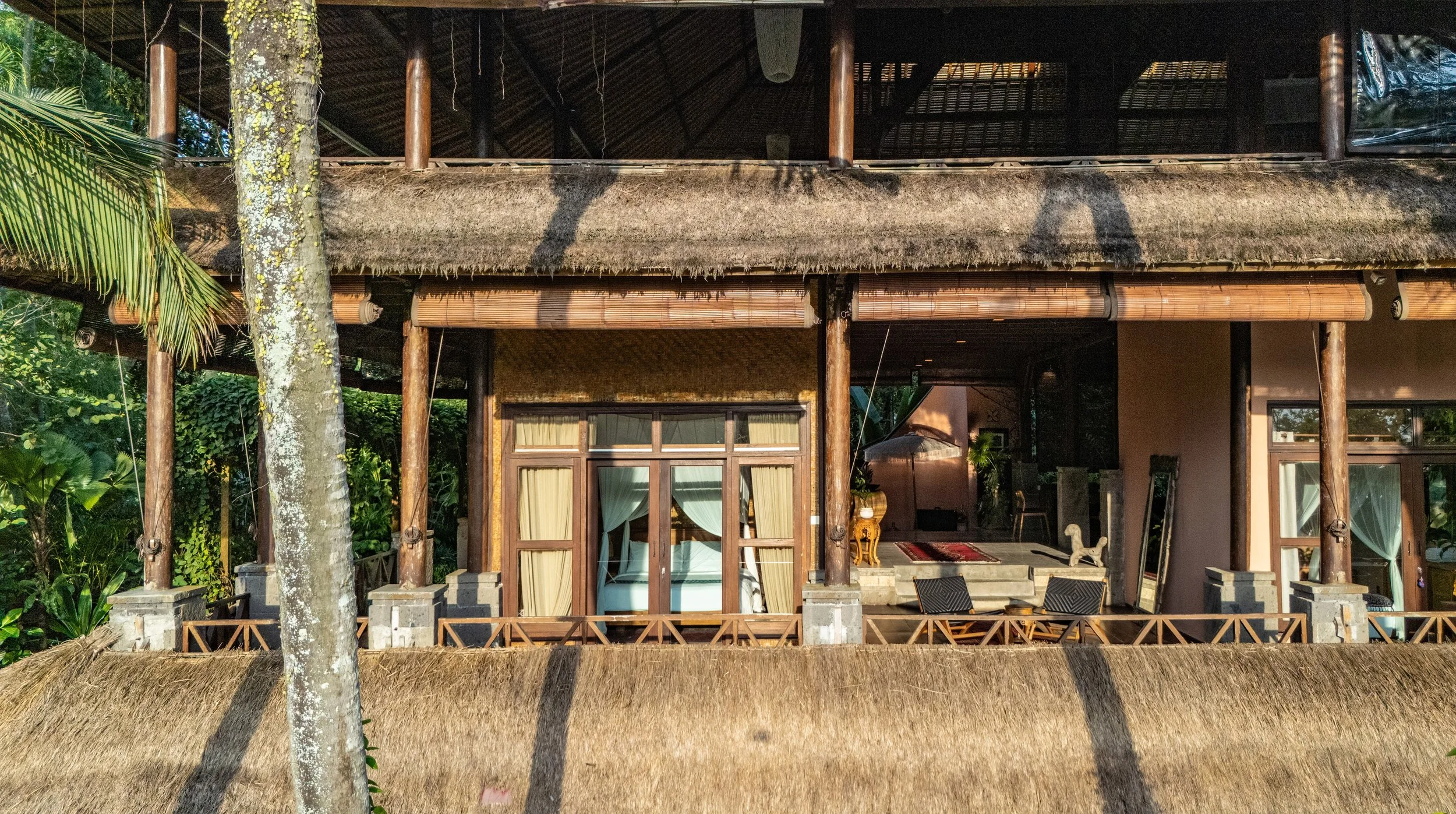 A two-story tropical house with a thatched roof, wooden columns, and large glass doors, surrounded by lush green plants and trees.