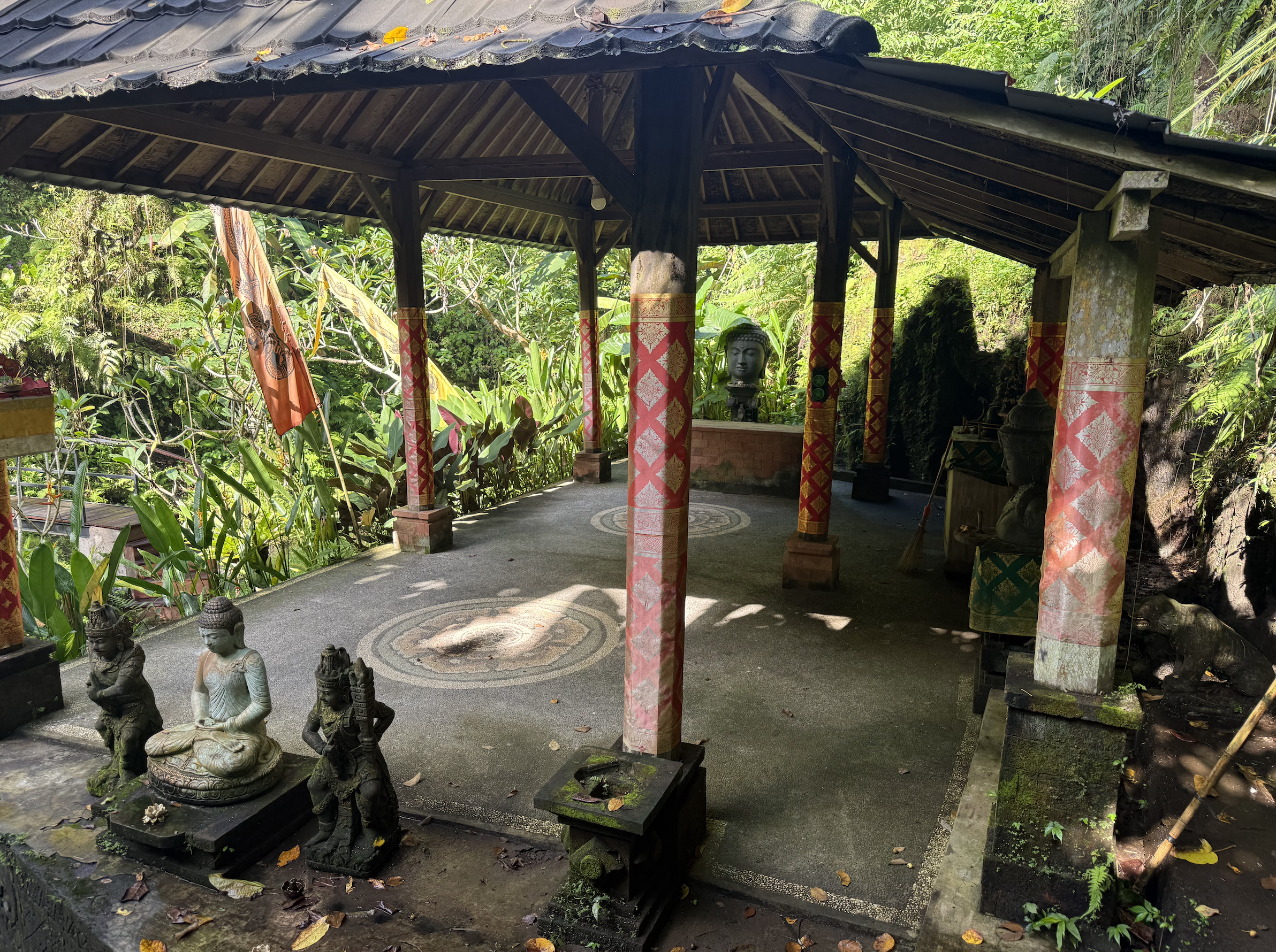 Small open-sided pavilion with a wooden roof and decorated columns in a lush green garden. Statues of seated figures in meditation are visible in the foreground, and a Buddha statue is seen inside the pavilion.High-end retreat center in Pejeng, Ubud