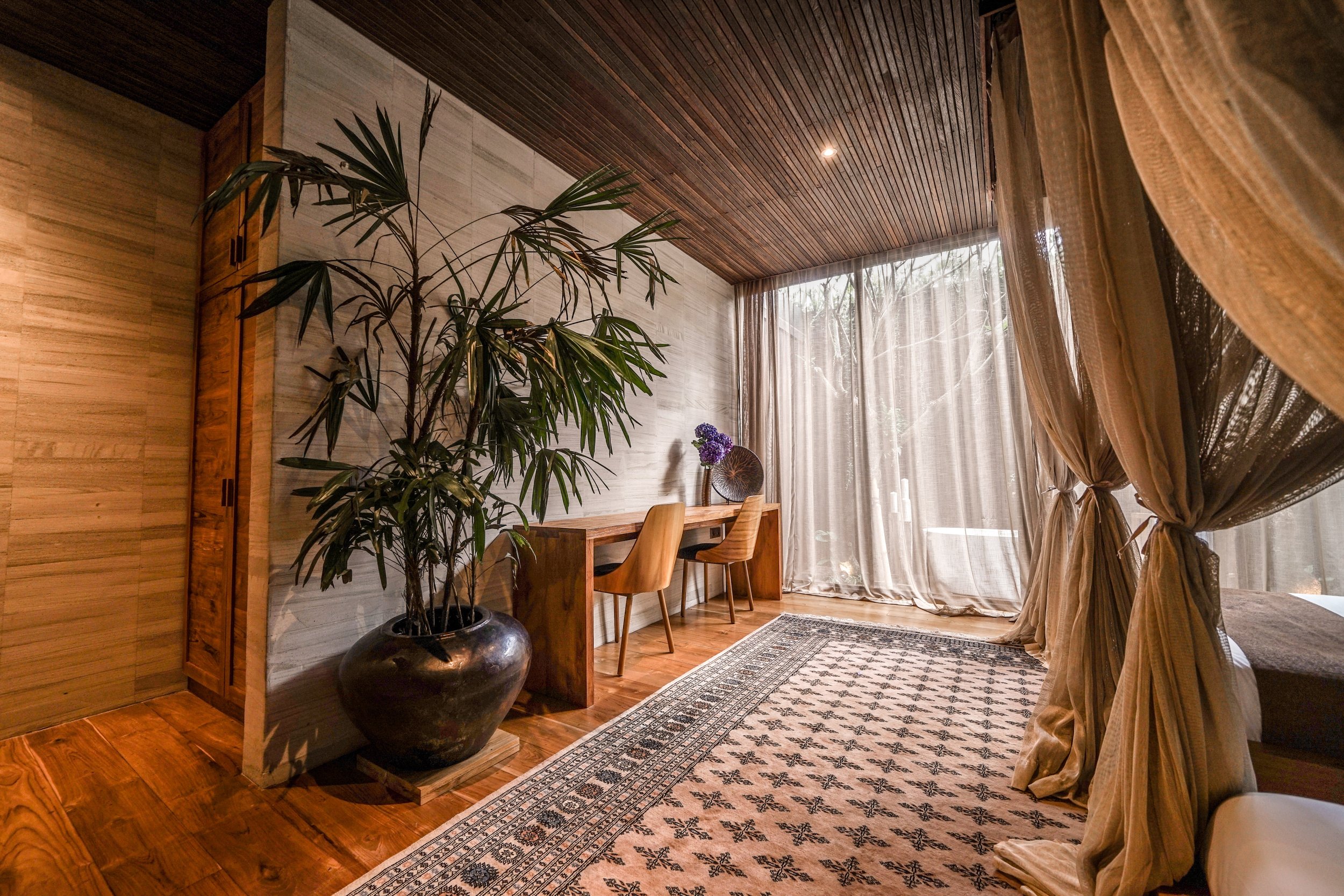 A corner of a room with wooden paneling, a large potted plant, a wooden dining table, chairs, a patterned rug, and large windows with sheer curtains. High-end retreat center in Pejeng, Ubud, Bali
