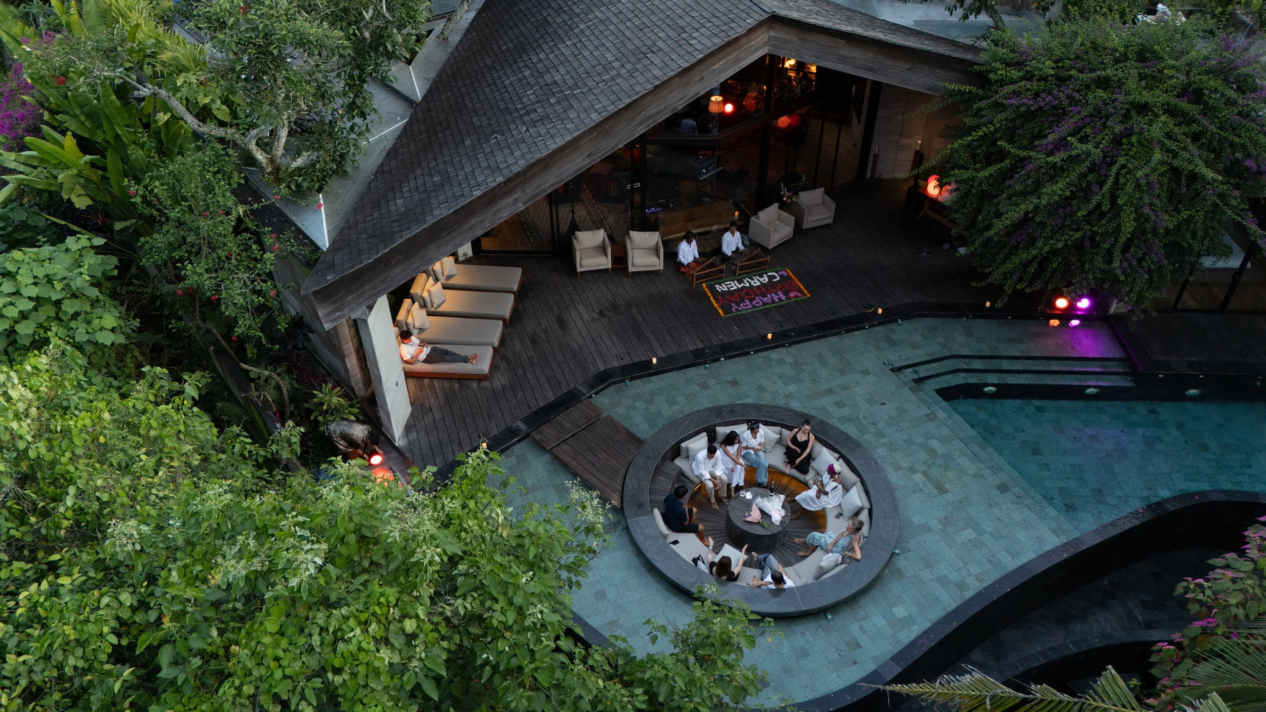 Aerial view of an outdoor hotel lounge area adjacent to a pool, with lounge chairs, a circular sofa, and several people sitting and relaxing, surrounded by lush greenery and trees. High-end retreat center in Pejeng, Ubud, Bali