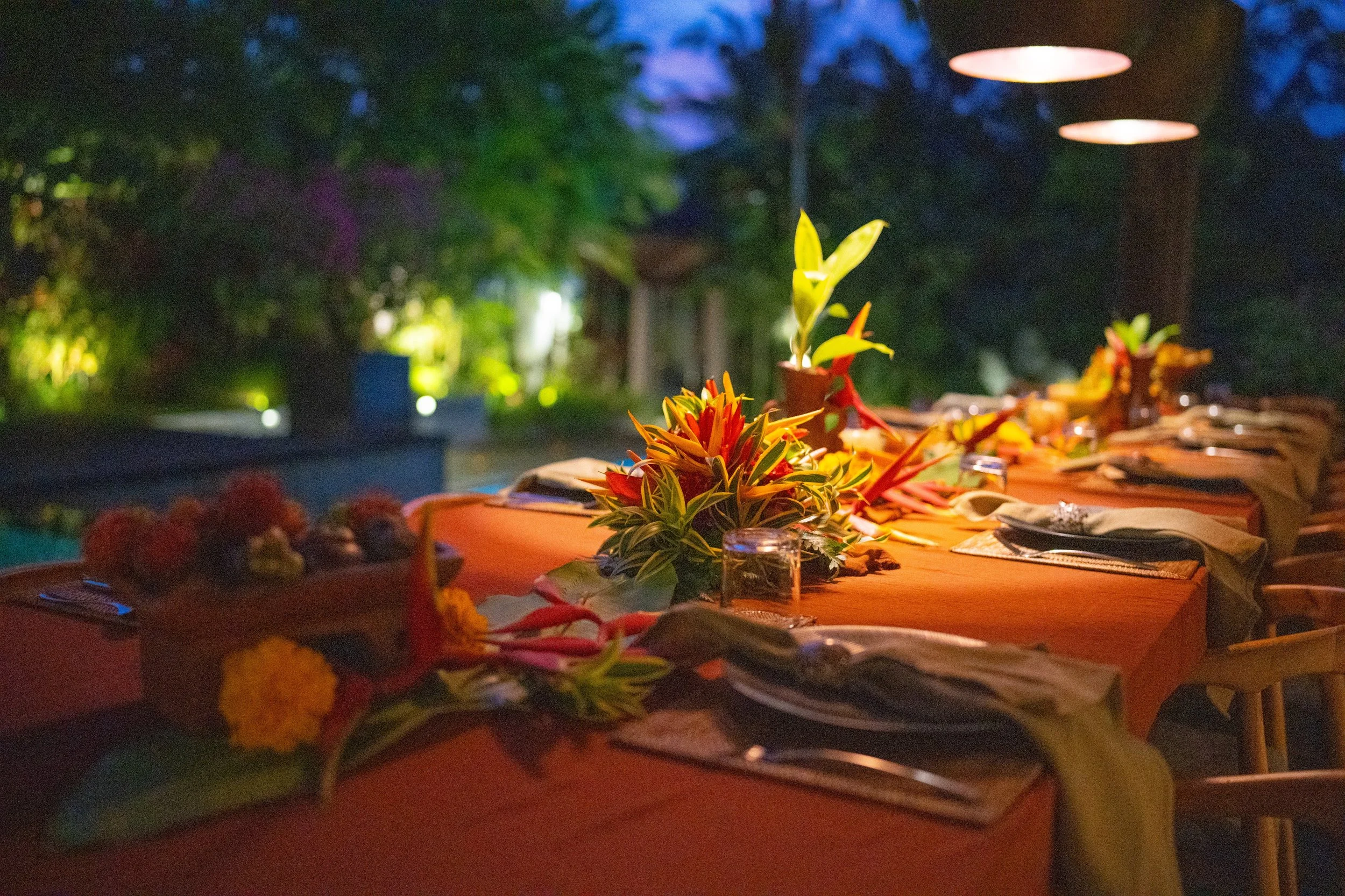 A table set for an outdoor dinner gathers with vibrant flower arrangements, surrounded by lush greenery, under evening lighting. High-end retreat center in Pejeng, Ubud, Bali