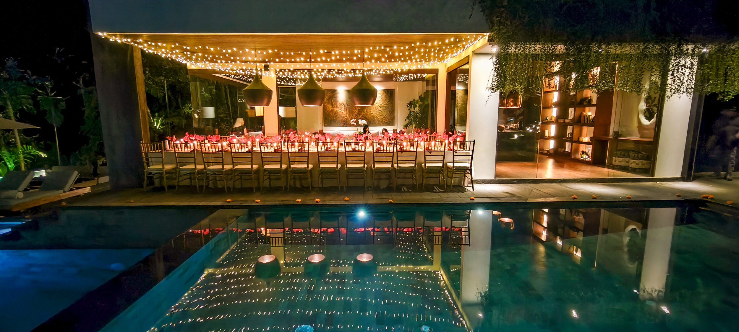 An outdoor dining area with a long table set for a special event, illuminated by string lights overhead, reflecting in a swimming pool in the foreground. High-end retreat center in Pejeng, Ubud, Bali