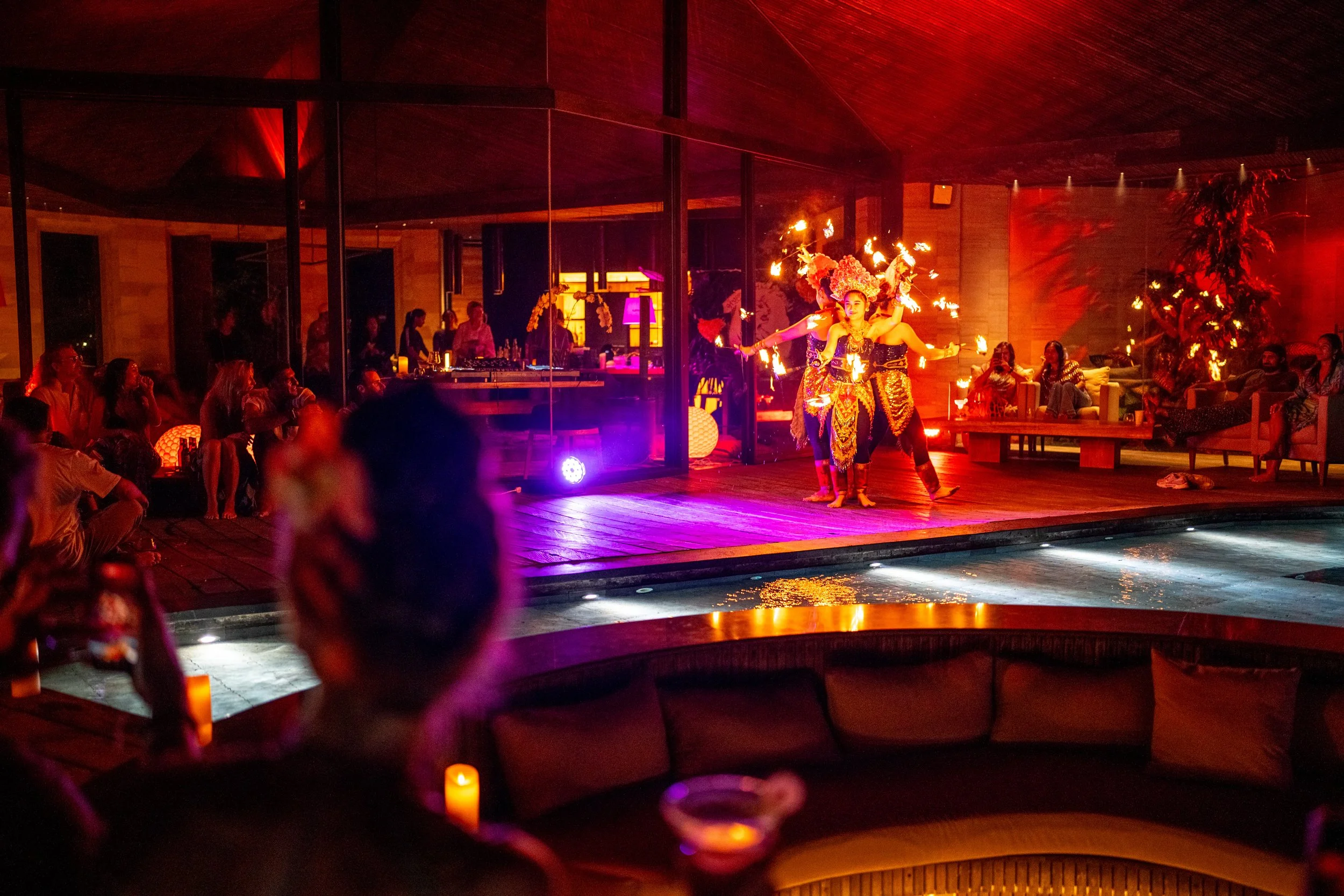 A nighttime scene of a bar or club with lounge seating, candles, and people watching a fire dance performance on stage. The performers are holding flaming torches and wearing colorful costumes.High-end retreat center in Pejeng, Ubud, Bali