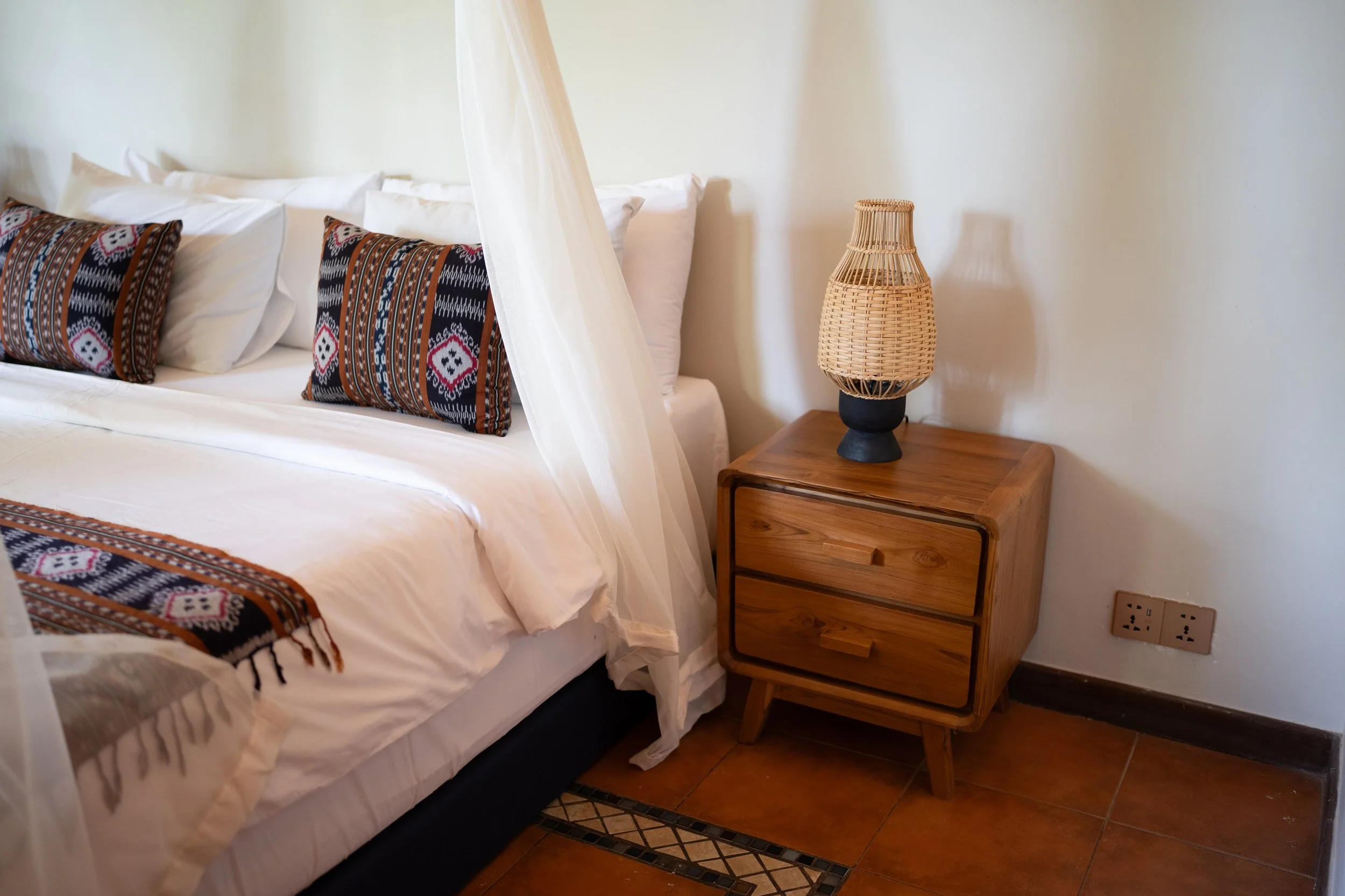 A cozy bedroom with a white bed featuring colorful patterned pillows, a wooden nightstand with a woven lamp, and a power outlet on the wall. High-end retreat center in Pejeng, Ubud, Bali