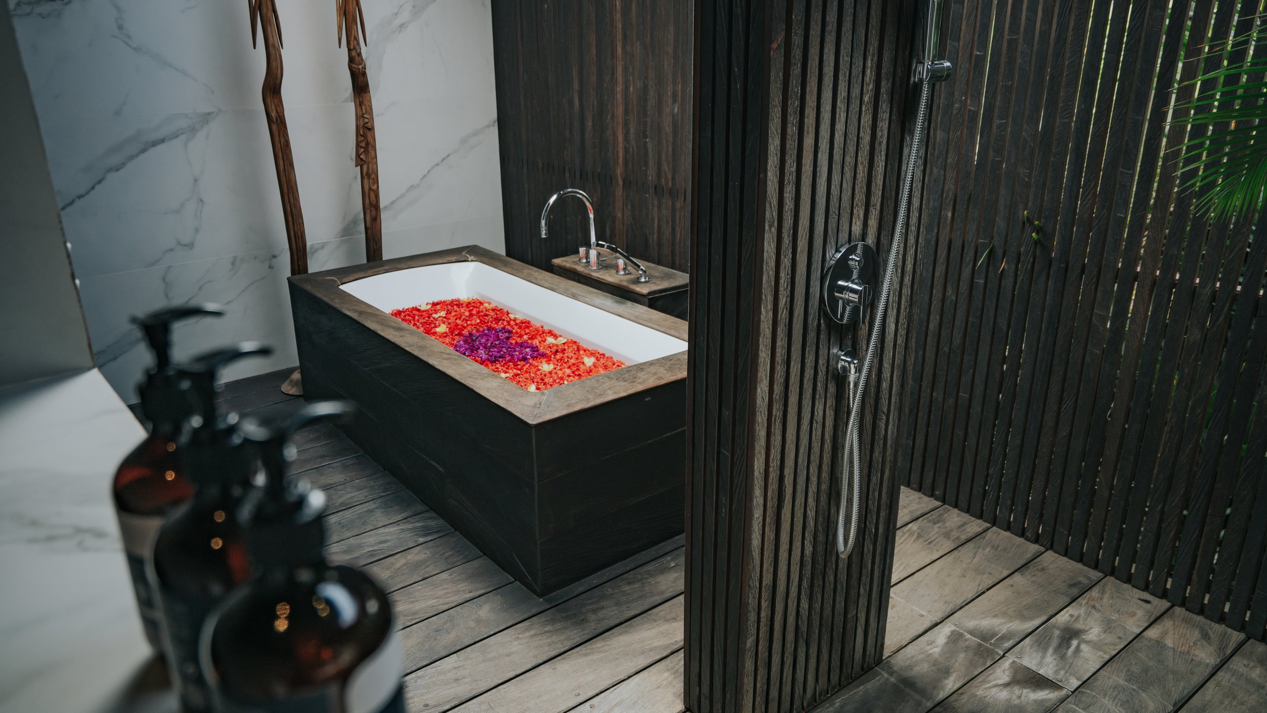 A bathroom with a black bathtub filled with orange and purple flower petals, a marble wall, and a wooden wall with a rainfall showerhead and a handheld shower attached.