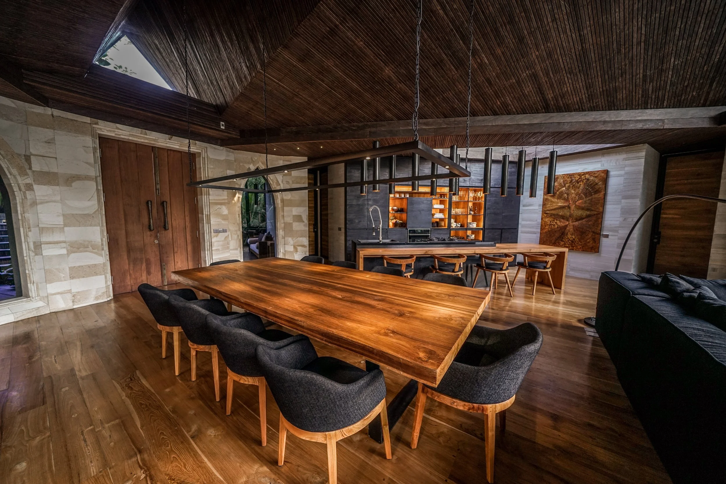 Modern dining room with a large wooden table surrounded by black upholstered chairs, a built-in kitchen with open shelves, and a slanted wooden ceiling with a skylight.High-end retreat center in Pejeng, Ubud, Bali