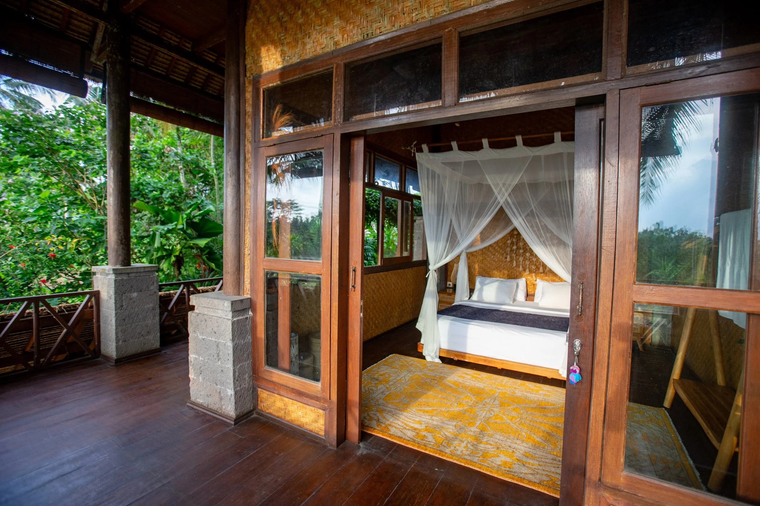 A tropical-style bungalow with an open wall, revealing a bedroom with a four-poster bed and mosquito net, surrounded by lush greenery and palm trees.