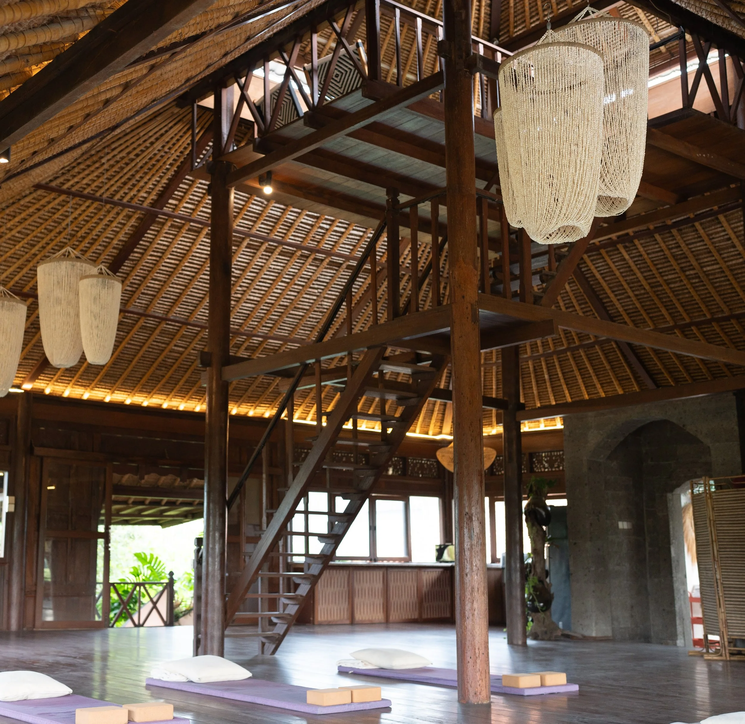 An interior view of a yoga or meditation space with purple mats, pillows, cork blocks, and white lampshades on the floor and a wooden staircase leading to a balcony with woven bamboo walls and a thatched roof ceiling. High-end retreat center in Pejen