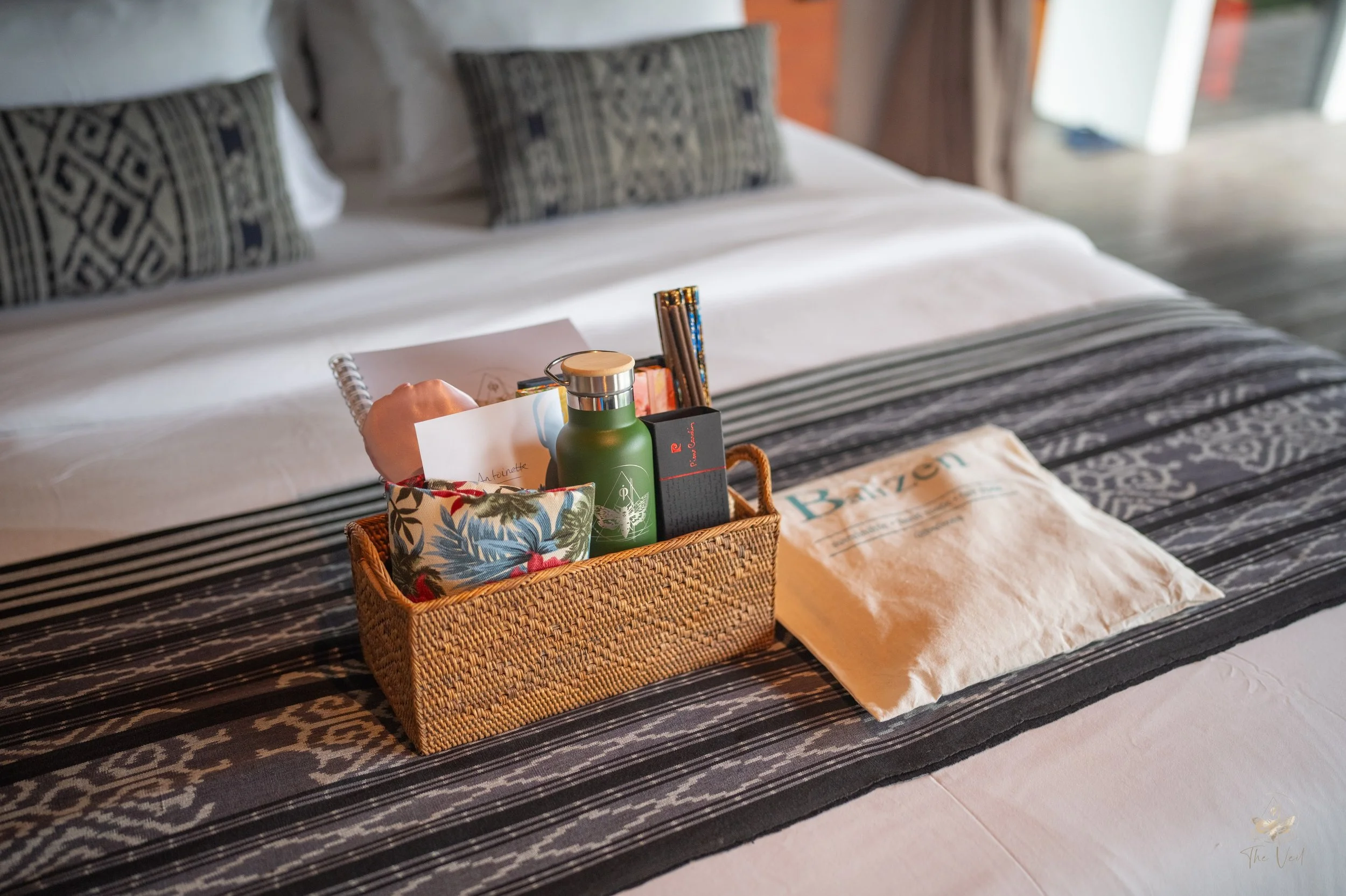 A basket on a hotel bed contains toiletries, a small notebook, and a colorful pouch, with a towel and pillows visible in the background. High-end retreat center in Pejeng, Ubud, Bali