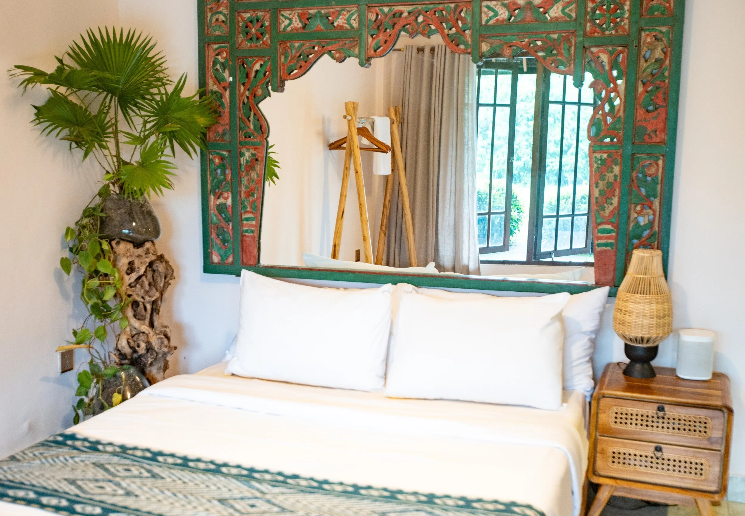 A living space with a large tropical plant on the left, a decorative mirror with intricate wood carvings in green and red, a window with beige curtains in the background. High-end retreat center in Pejeng, Ubud, Bali