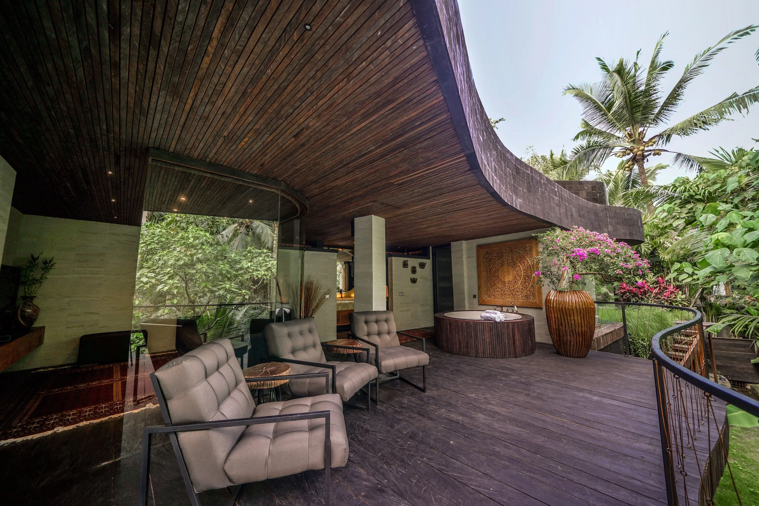 Outdoor lounge area with beige chairs, tropical plants, and a wooden ceiling under a curved roof, with lush greenery and palm trees in the background.