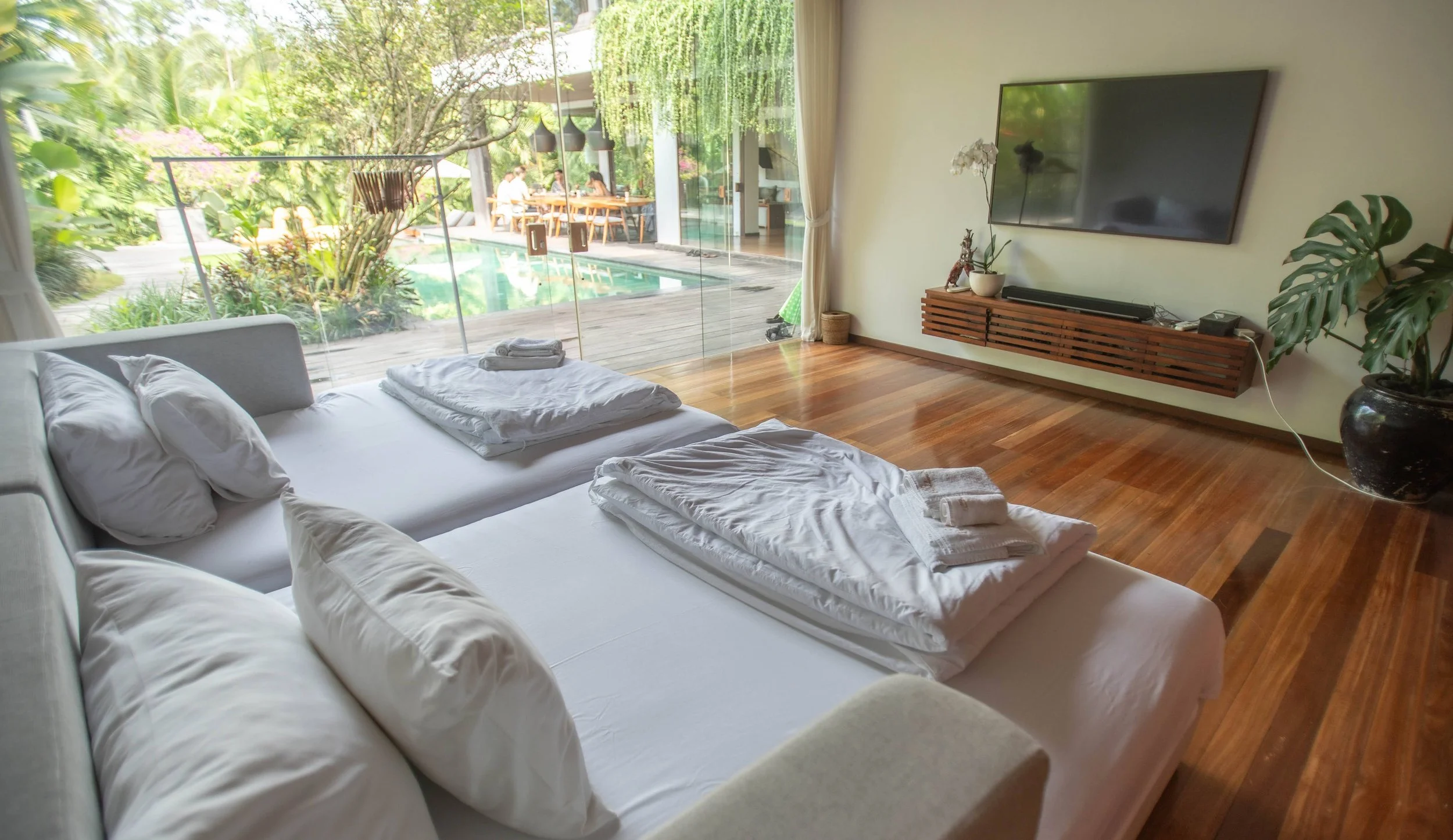 A modern bedroom with a large white bed, neatly made with white pillows and towels, facing a glass wall with a view of a lush garden and outdoor dining area. High-end retreat center in Pejeng, Ubud, Bali