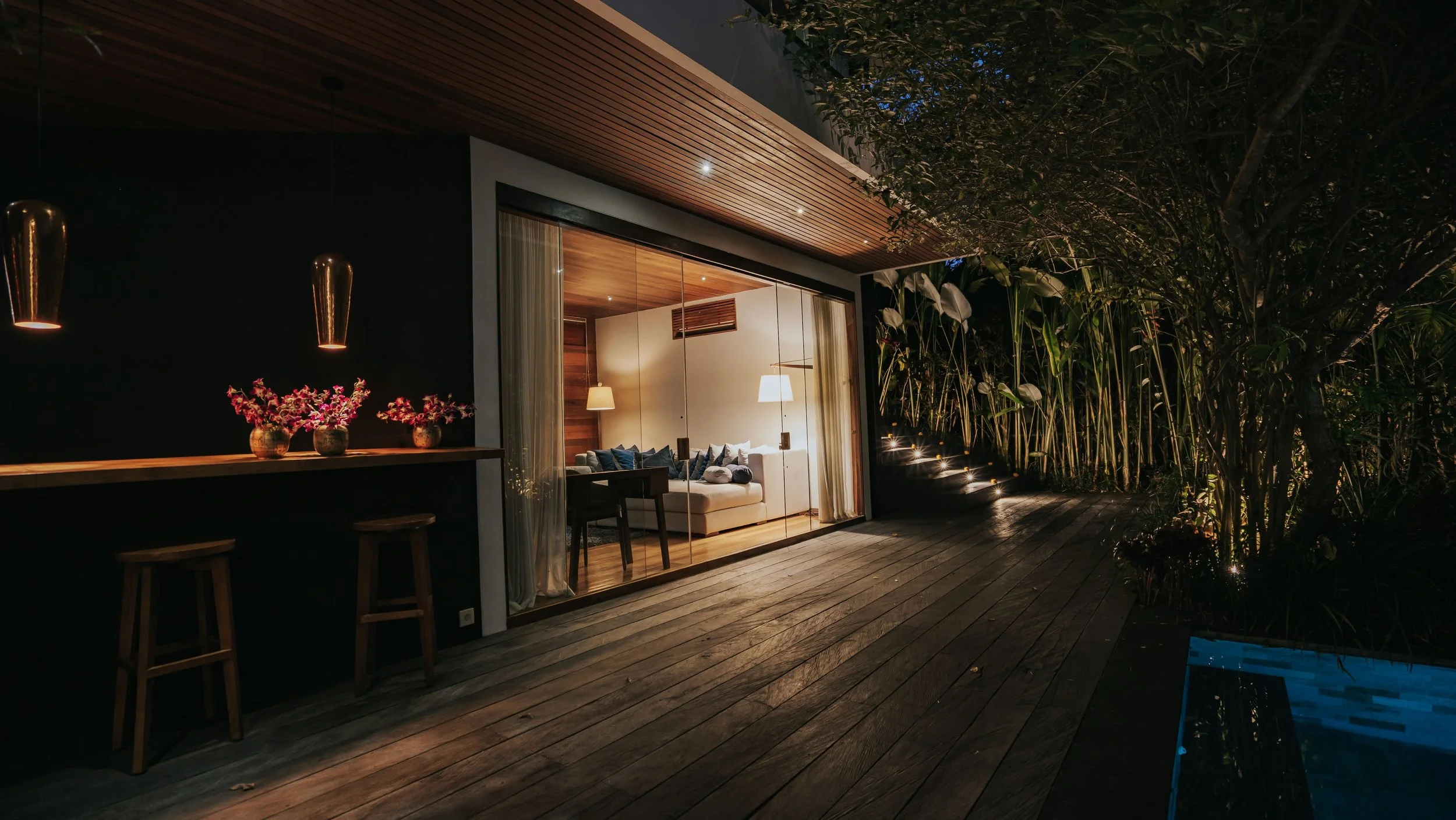 Modern house exterior at night with large glass sliding doors, cozy interior lit with lamps, wooden ceiling and floor, outdoor patio with chairs, plants, and trees illuminated by pathway lights. High-end retreat center in Pejeng, Ubud, Bali