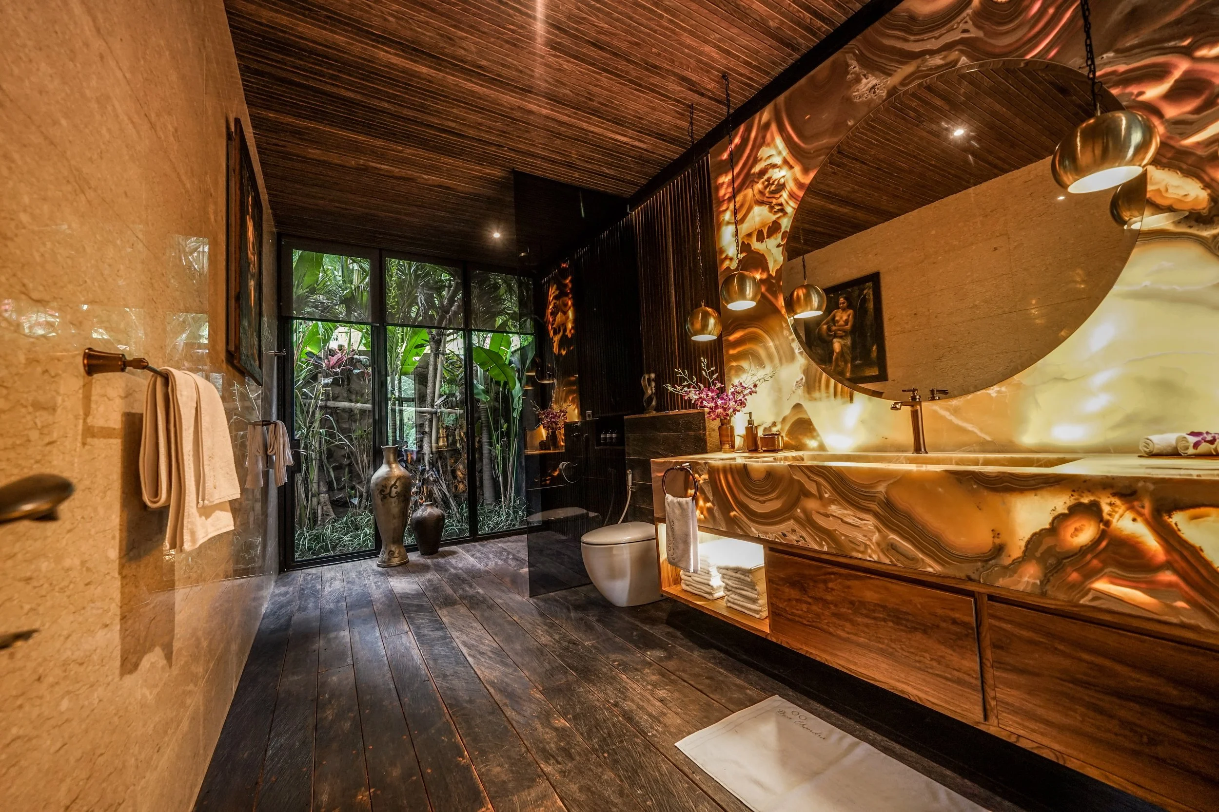 Elegant bathroom featuring a large marble vanity with warm lighting, wood-paneled ceiling, large window with greenery outside, and decorative vases. High-end retreat center in Pejeng, Ubud, Bali