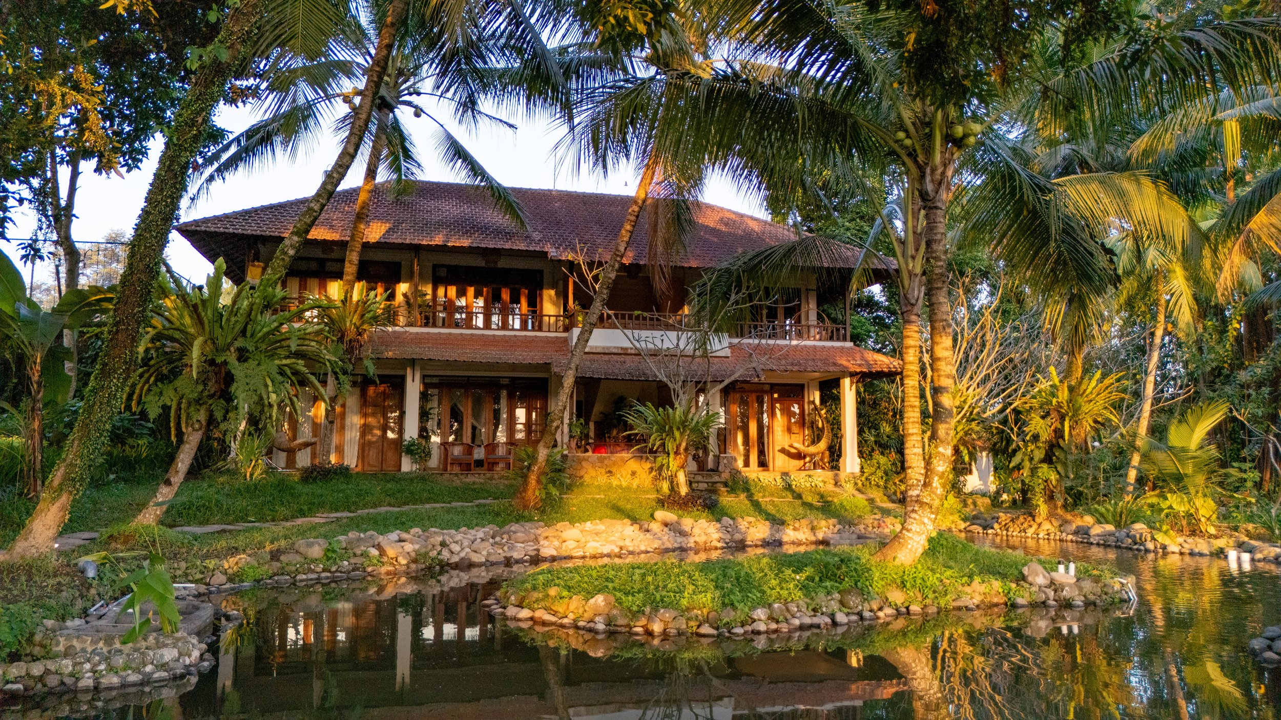 A house surrounded by palm trees and lush greenery, with its lights on at sunset, reflecting in a small pond in the foreground. High-end retreat center in Pejeng, Ubud, Bali