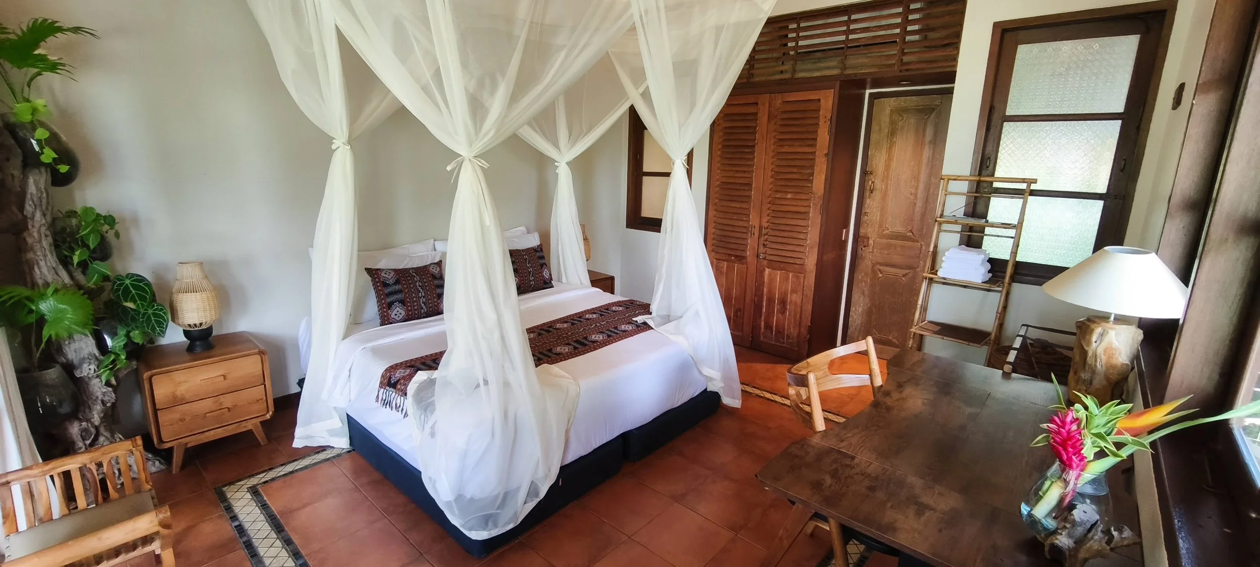 A cozy bedroom with a four-poster bed draped in white sheer curtains, two patterned pillows, and a quilted bed runner. The room features wooden furniture, including a wardrobe, small bedside tables, and a writing desk. There are green plants, a table lamp, and a vase with tropical flowers near the window, which has frosted panes.