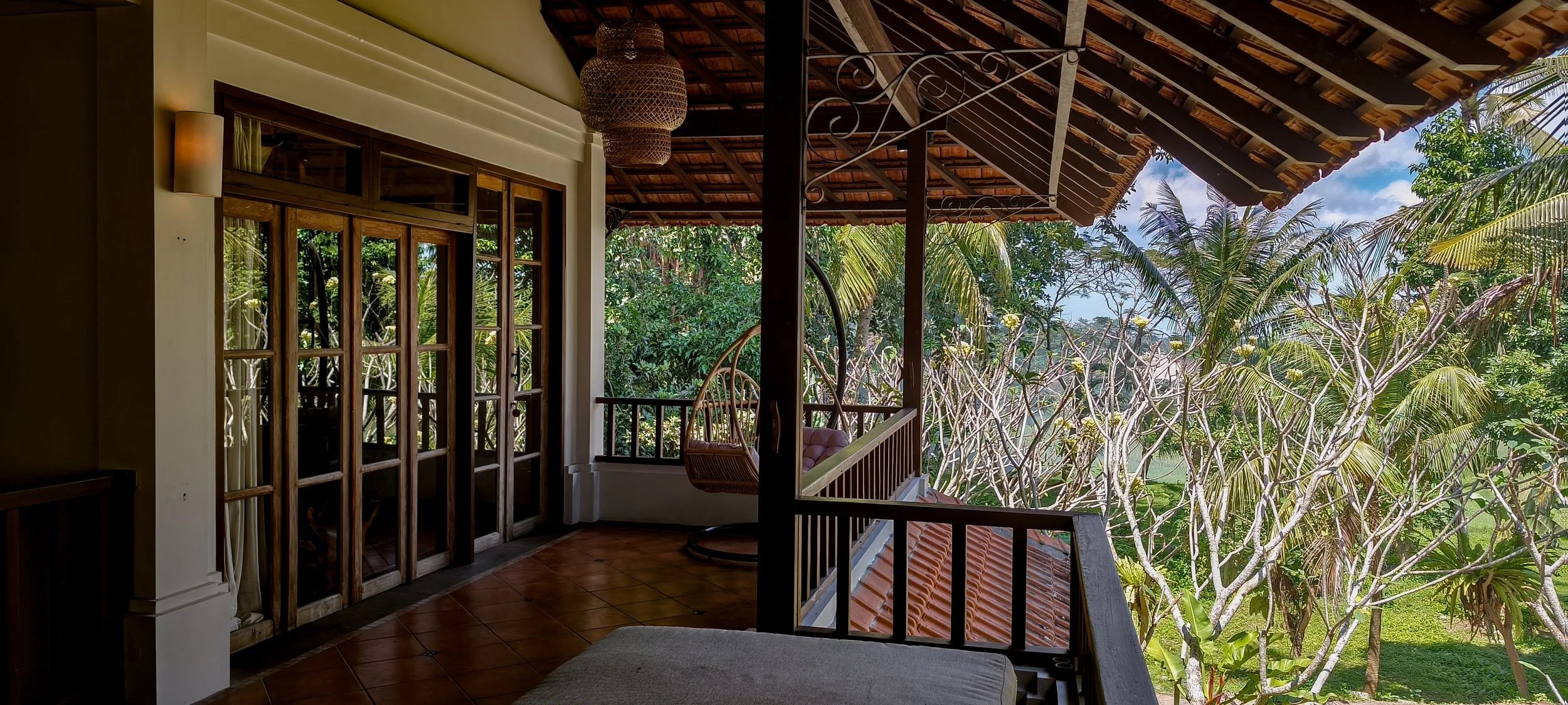A balcony with wooden doors, wicker hanging chair, and tropical trees outside on a sunny day.High-end retreat center in Pejeng, Ubud, Bali