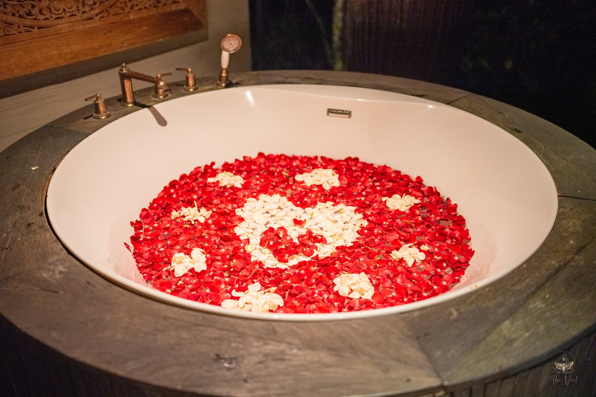 A bathtub filled with red rose petals and white flower petals, with a bronze faucet and handles, in a dimly lit bathroom.