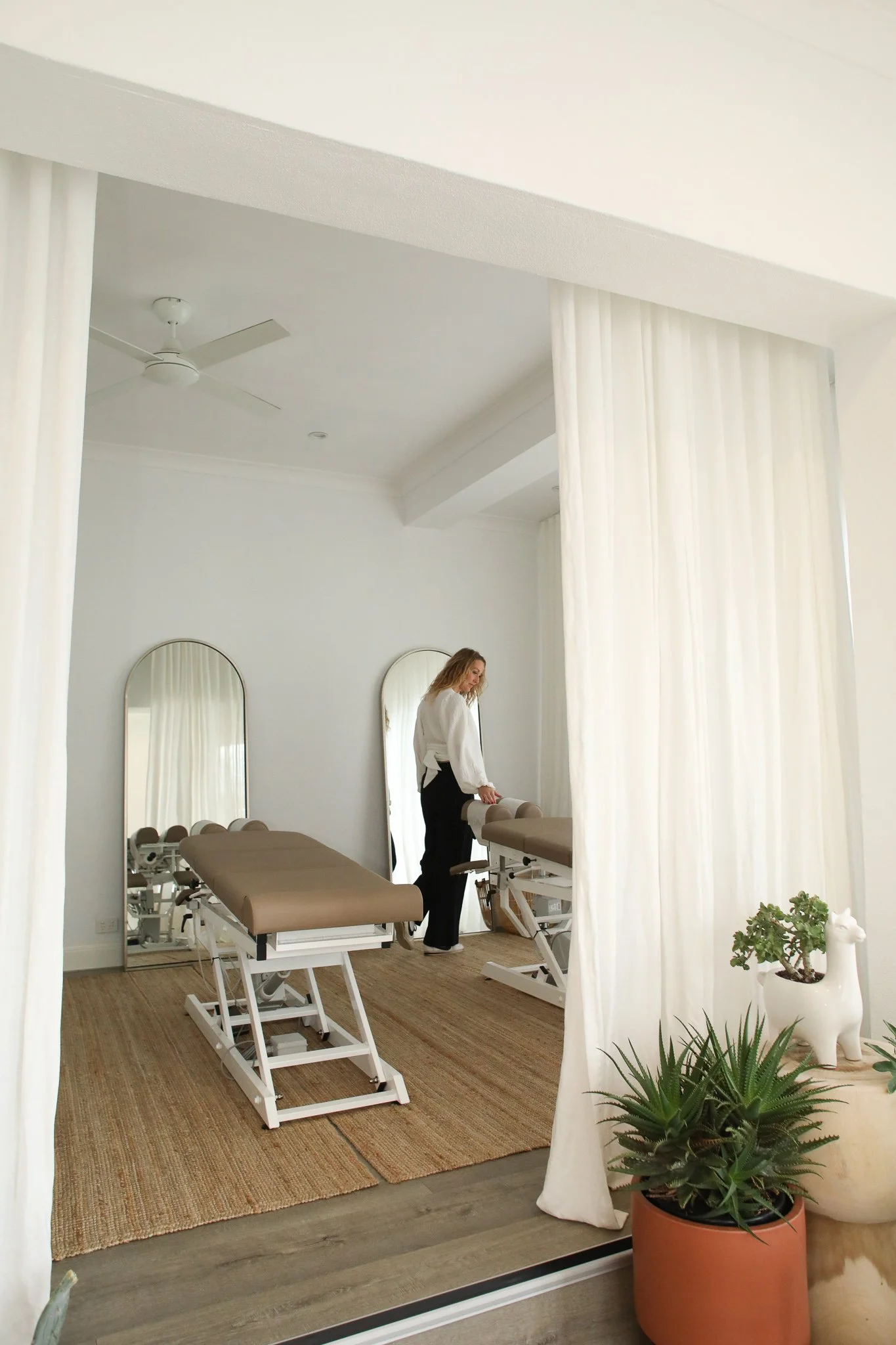 A woman in a white blouse and black pants standing inside a tranquil treatment room with two treatment beds, mirrors, floor-length white curtains, green plants, and a woven rug on a wood floor.