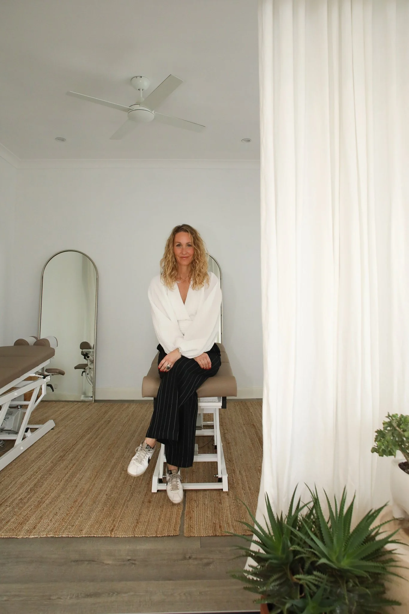 A woman with curly blonde hair, wearing a white blouse and black pinstripe pants, sitting on an examination table in a medical office, smiling at the camera. Behind her are two mirrors, a ceiling fan, and white curtains, with a plant in the foreground.