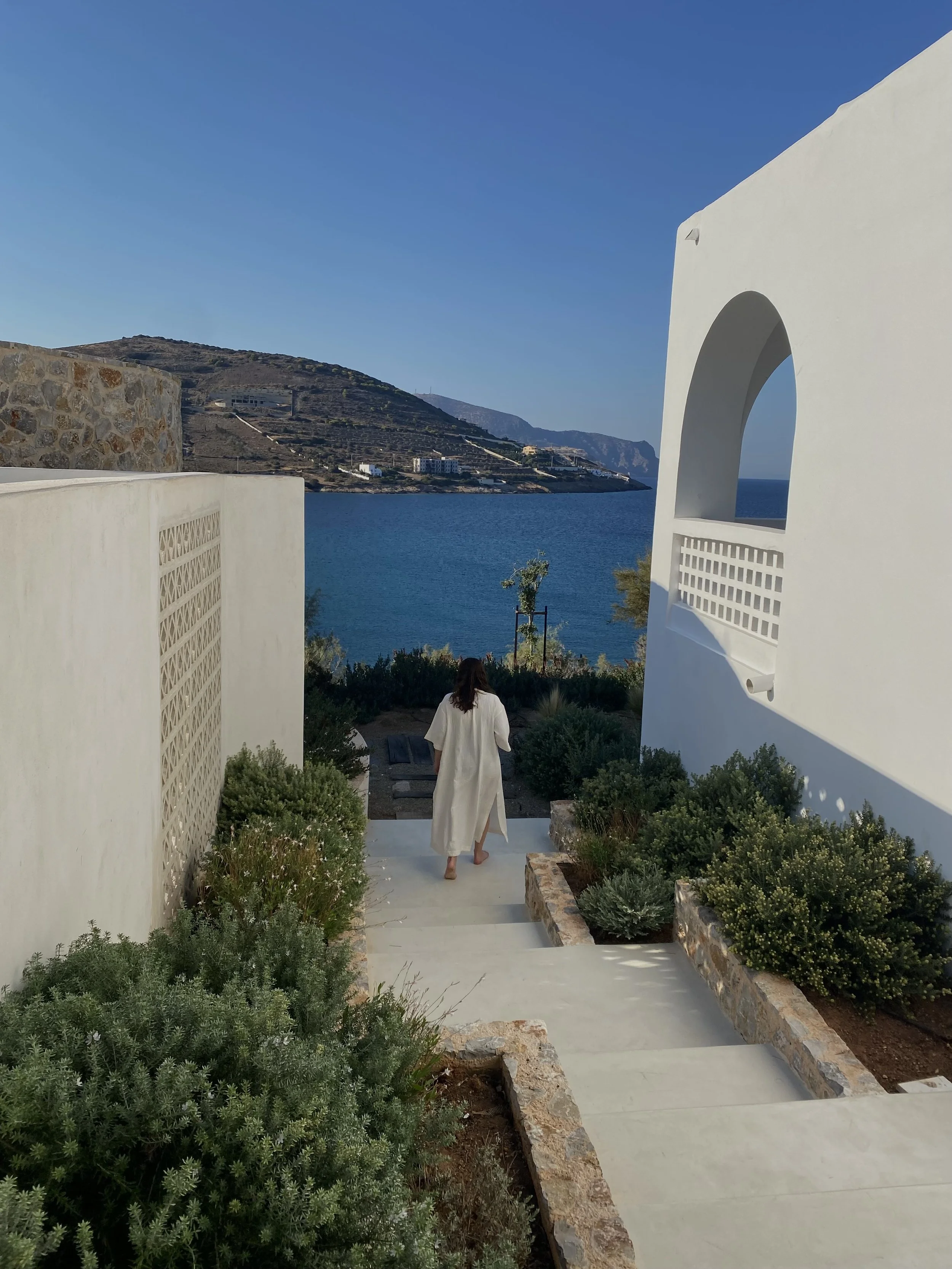 A woman in a white dress walking down a paved pathway with greenery on both sides, overlooking a large body of water with hills and buildings in the distance under a clear blue sky.