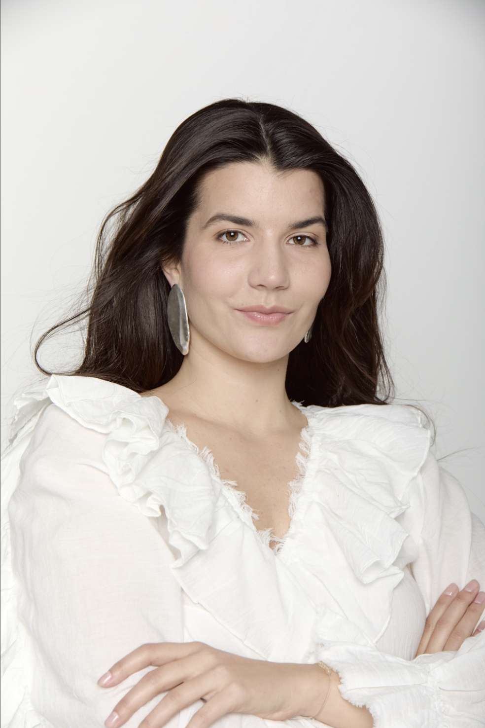 Woman with long dark hair standing confidently with a soft smile, arms crossed. She is wearing a white flowy blouse and silver statement earrings.