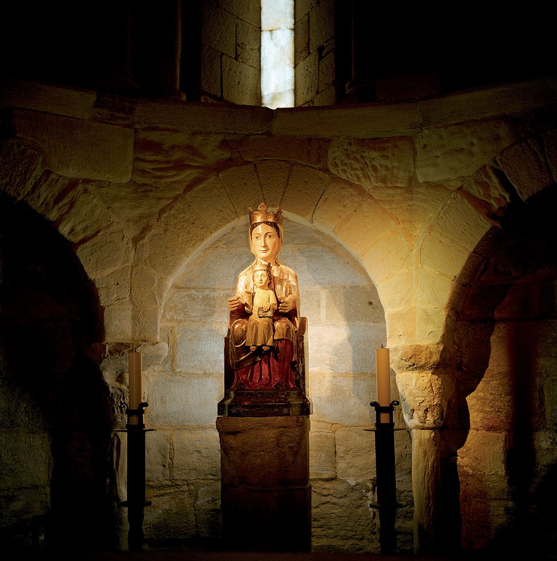 Religious statue of the Virgin Mary holding the Infant Jesus, set in a stone niche in a church or religious place, softly lit. Two candles are placed on either side of the statue.