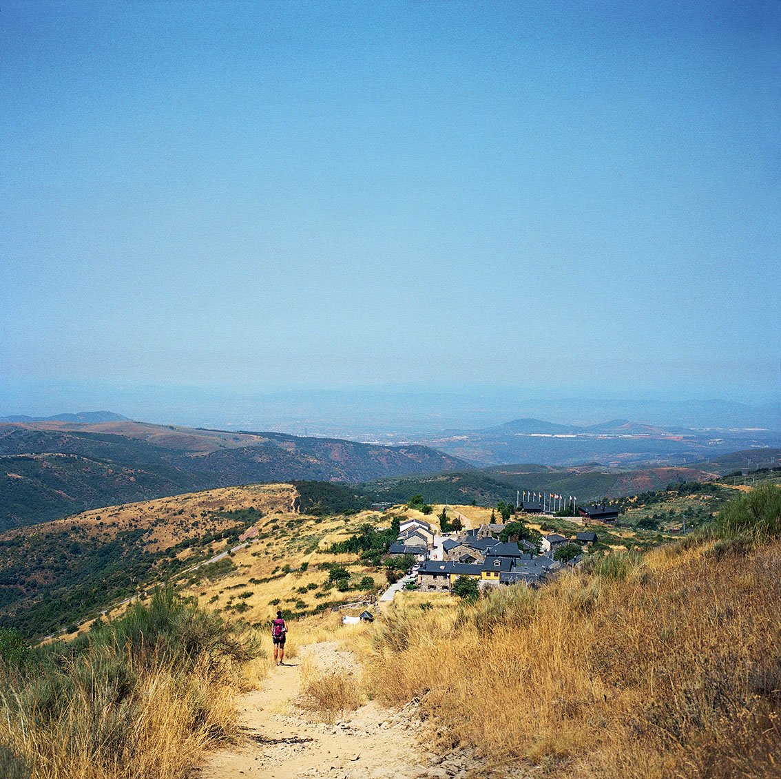 Hiker walking along a mountain path on a sunny day, view of a village of houses in the background, in a landscape of hills and valleys.