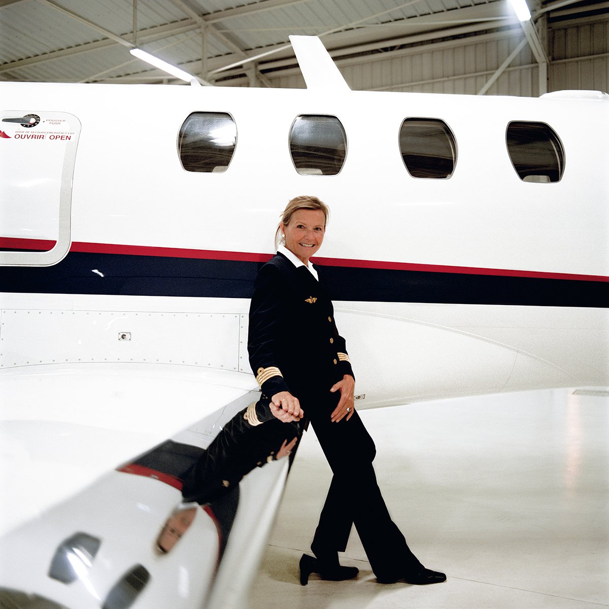A woman in a pilot's uniform poses next to a private plane inside a hangar.