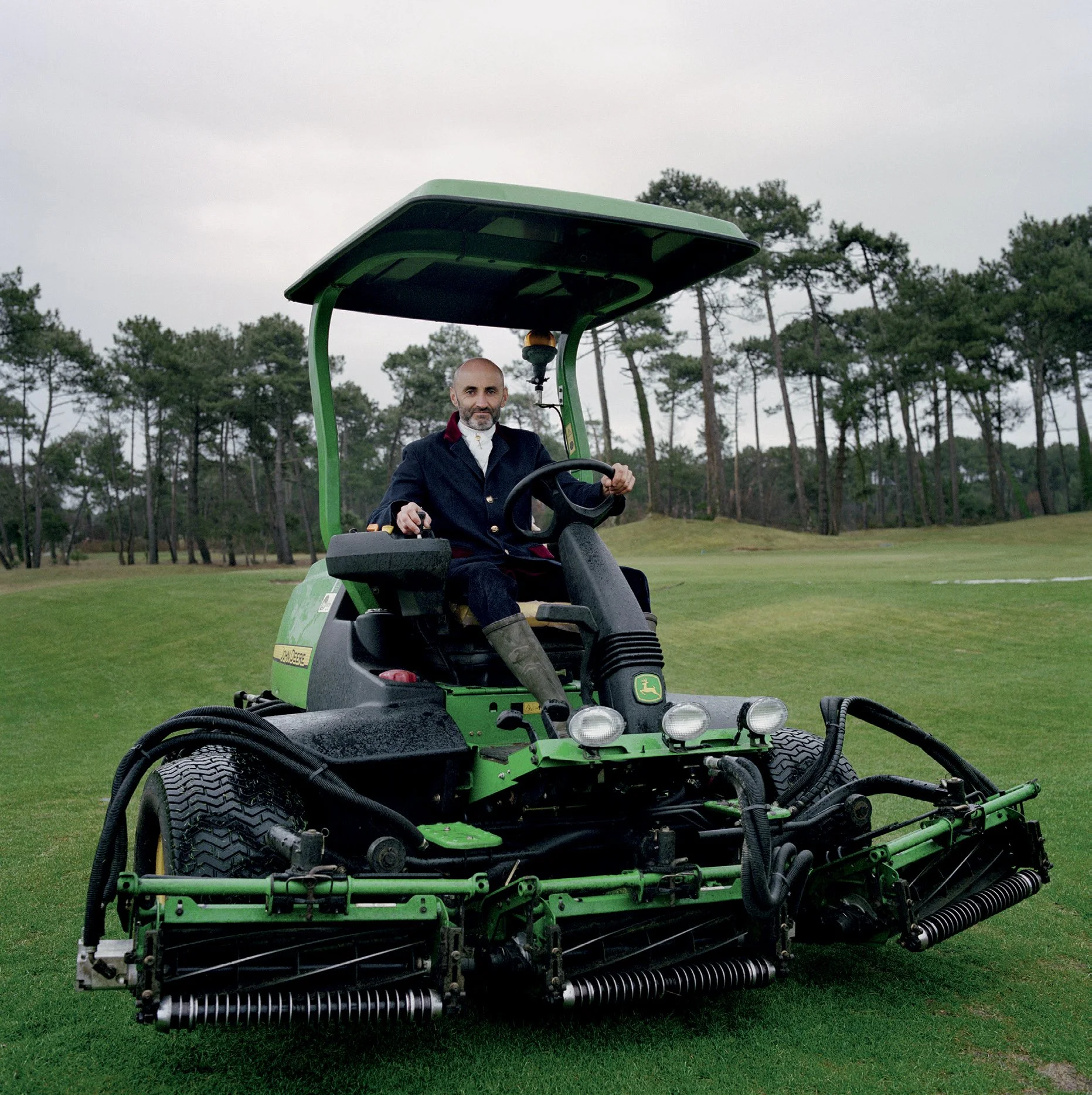 Man sitting on a green golf tonts machine on a golf course with trees in the background.