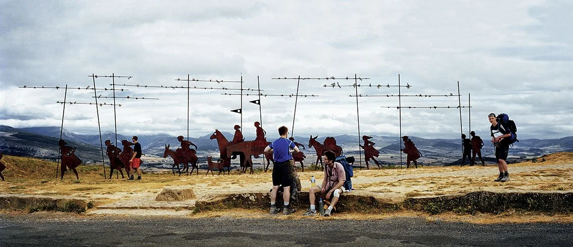 Group of tourists in front of an art installation depicting metal horses and human figures, outdoors in a rural landscape with mountains in the background.
