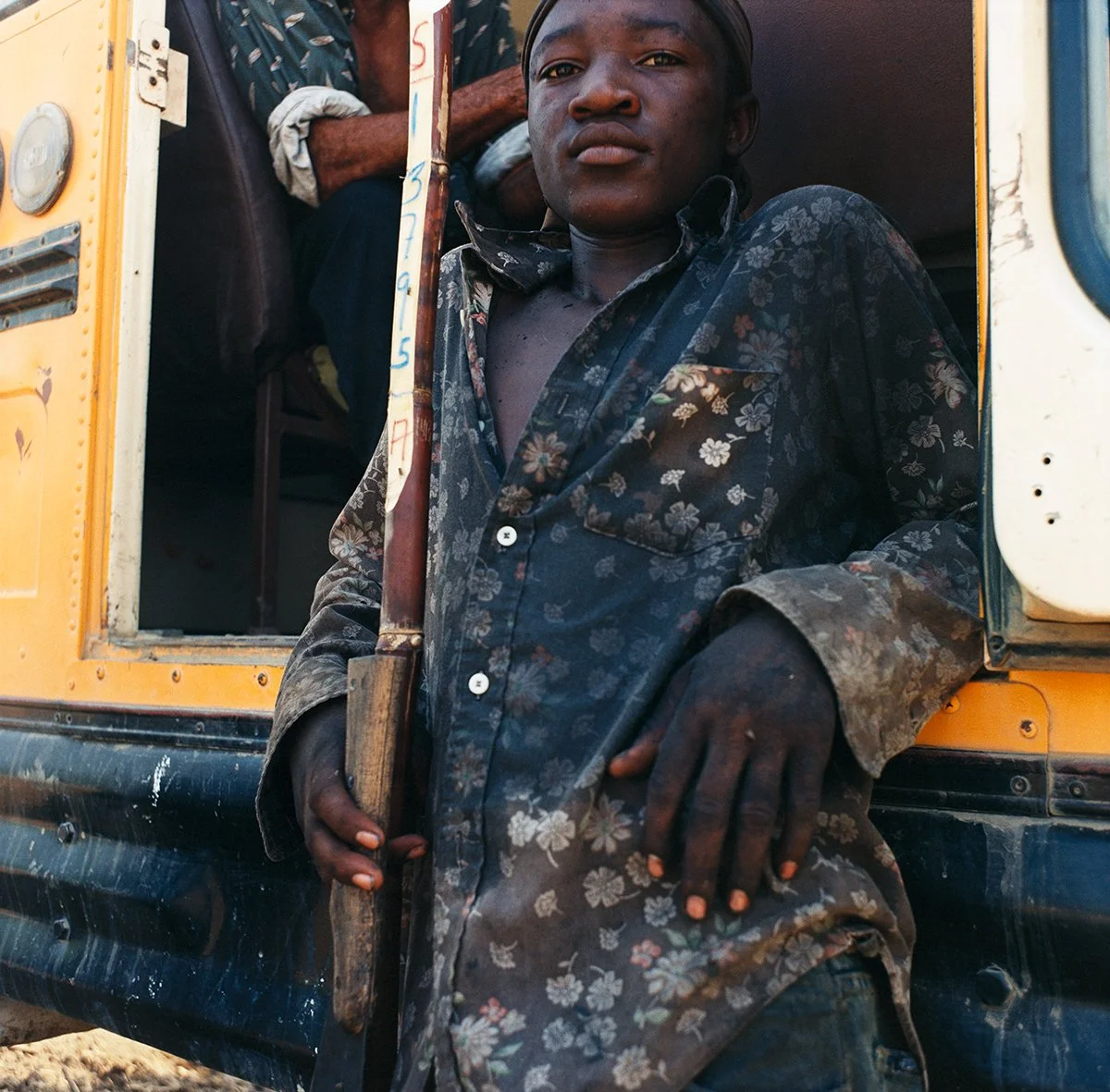 A young man poses outside a yellow vehicle, wearing a dark floral shirt and holding a wooden gun. He looks into the camera with a serious expression.