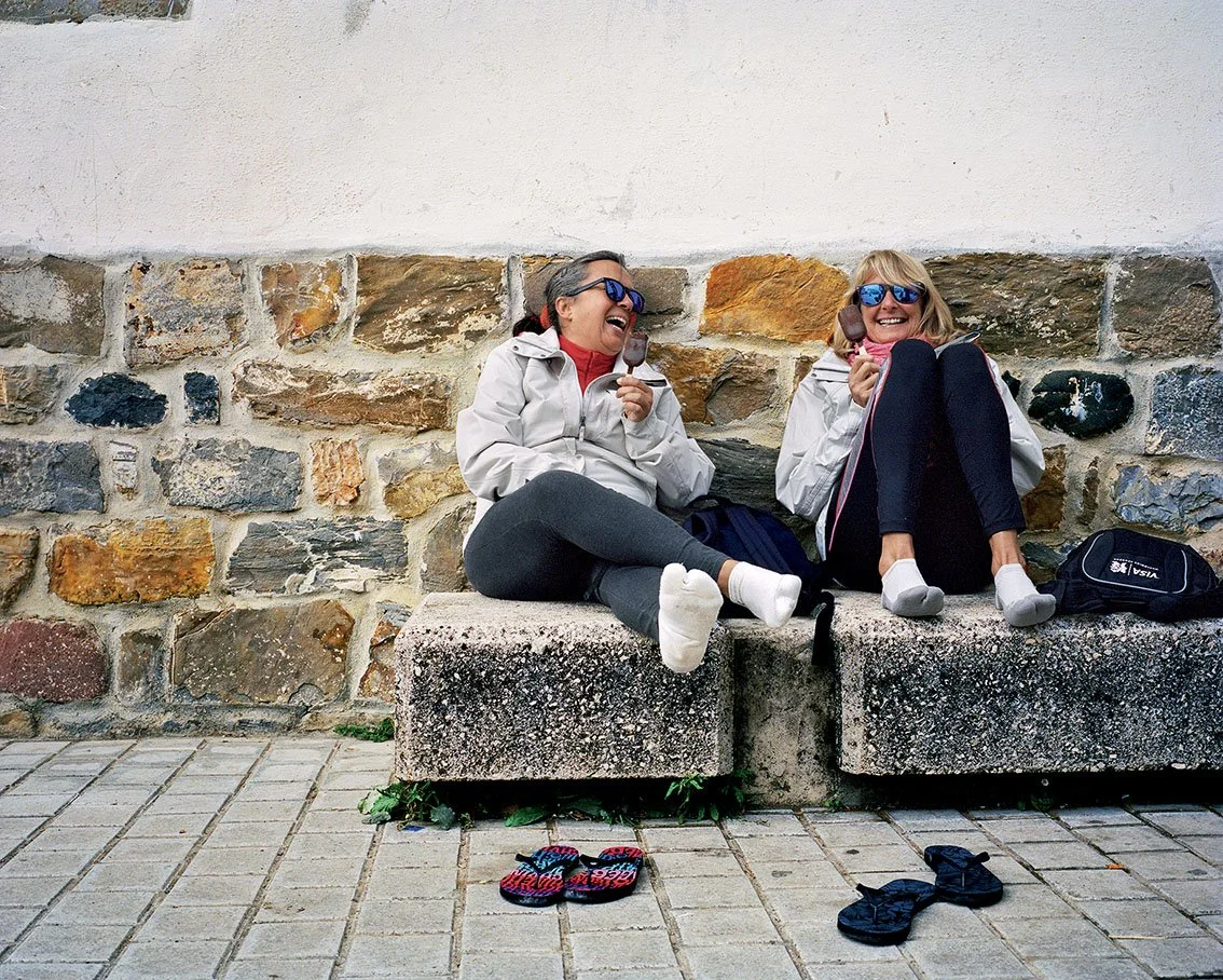 Two women sitting on a stone bench against a stone wall, laughing and enjoying an ice cream. One wears blue sunglasses and a gray coat, the other mirrored sunglasses and a white coat, with sandals and shoes.