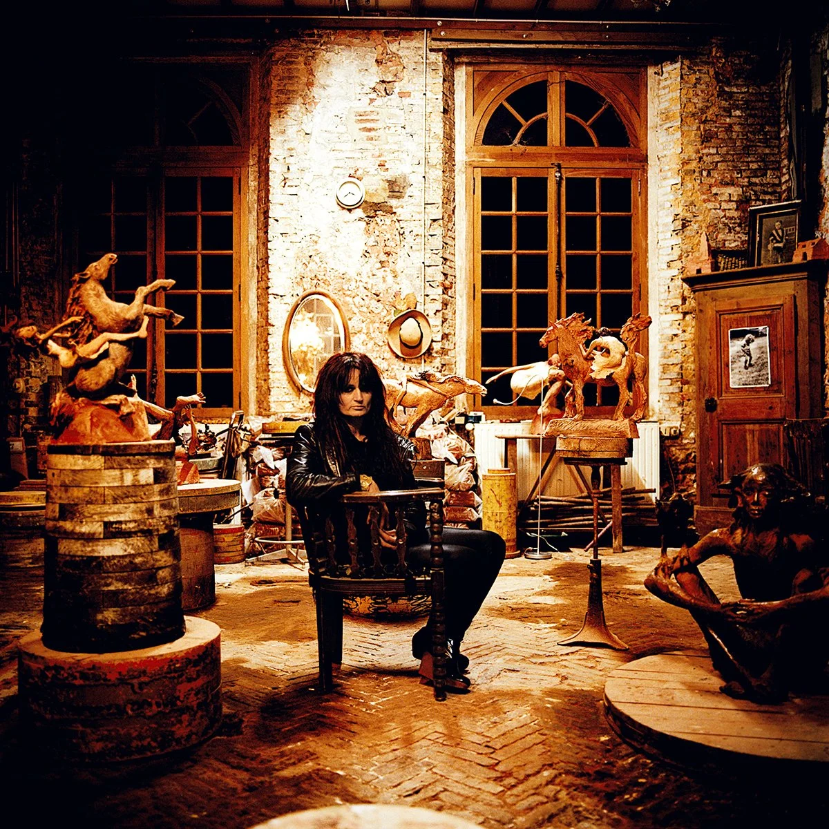 A female artist sits in an art studio, surrounded by wooden sculptures and works of art in a space with brick walls, large wooden windows and various objects.