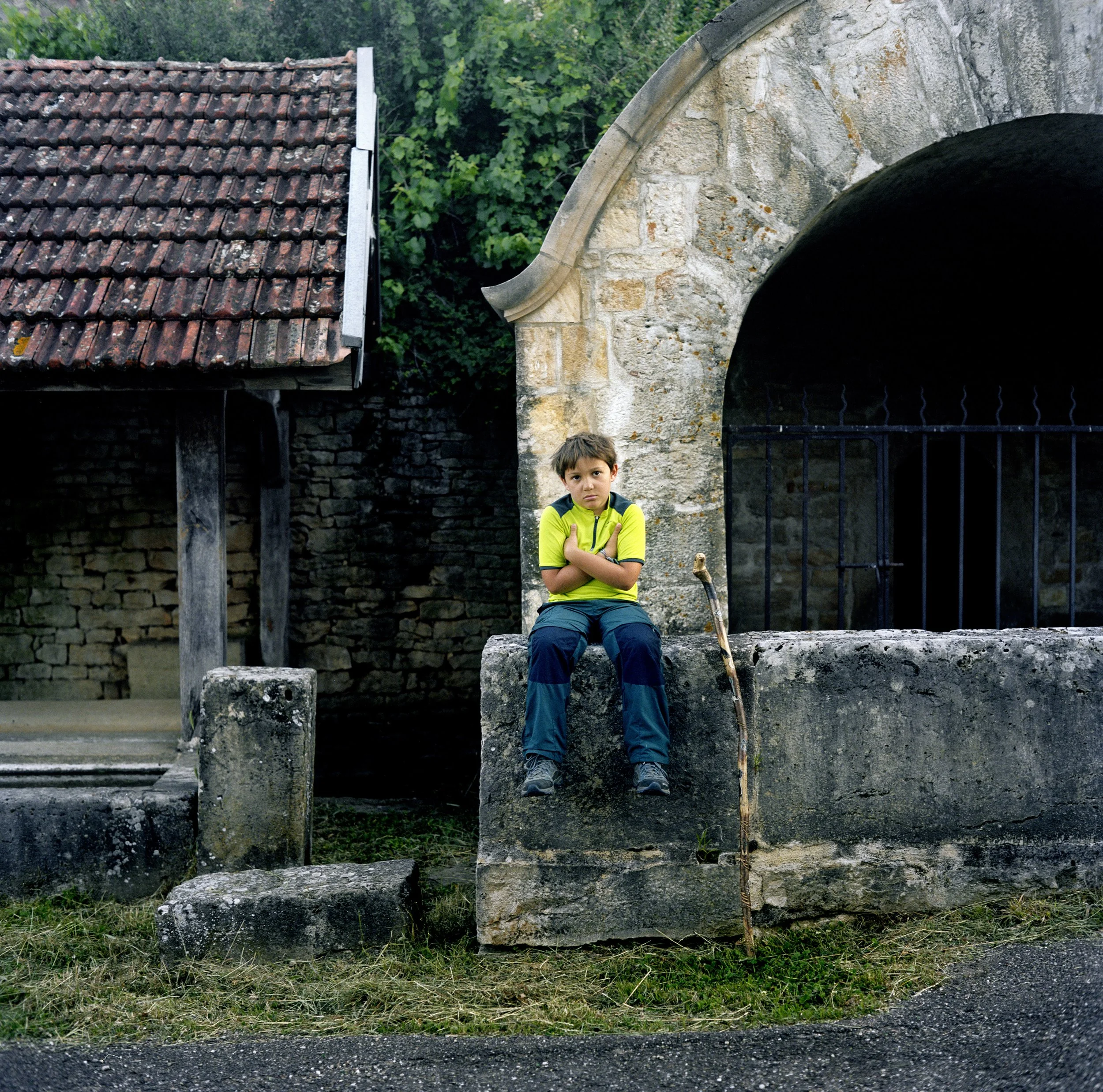 A boy sitting on a low stone wall, next to an old stone structure with a building in the background, in a rural setting with green trees.