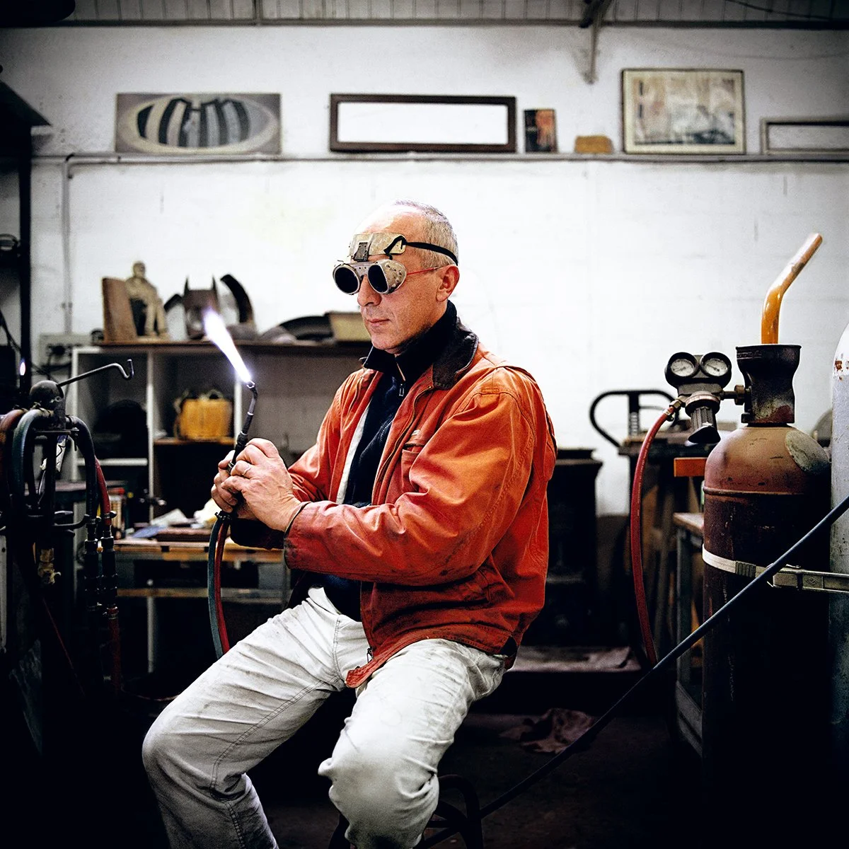 A man wearing goggles and an orange coat uses a lighter in a welding shop, surrounded by welding equipment and tools.
