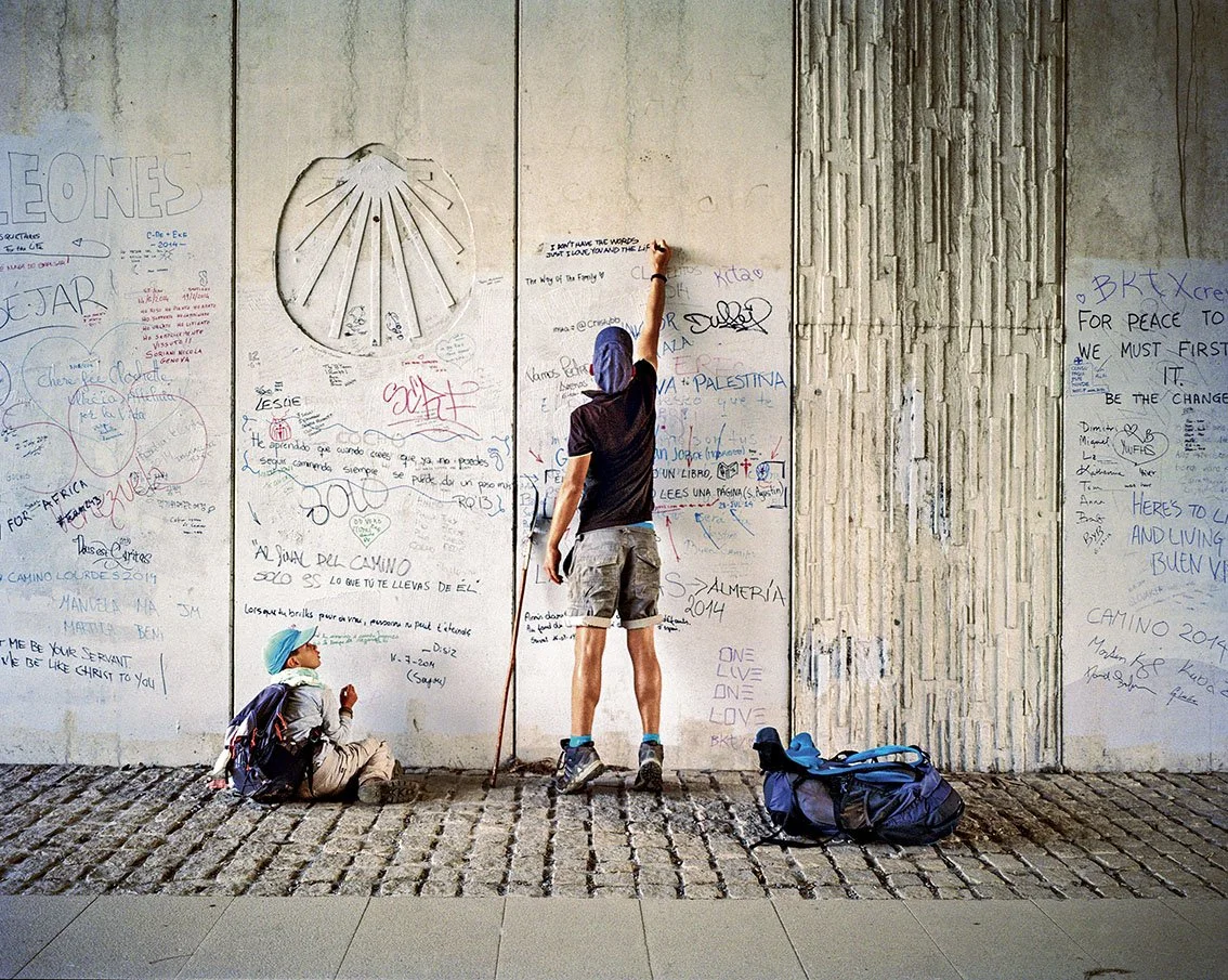 Two people, one standing and one sitting, in front of a wall lined with graffiti and written messages, one person writing on the wall. The man is wearing a black T-shirt, grey shorts and a blue beanie, with a backpack beside him. The person has