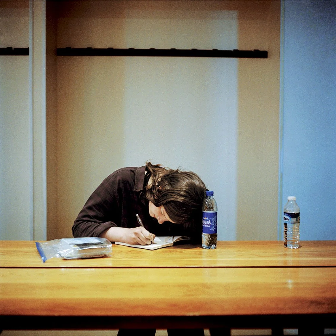 A woman sitting at a wooden table, writing in a notebook, with two bottles of water in front of her.