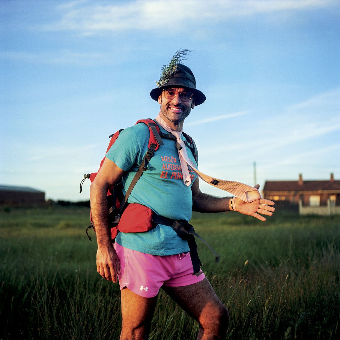 Smiling man with a hat decorated with vegetation, wearing a backpack, a blue t-shirt with red writing, pink shorts and sunglasses, in a field during sunset.